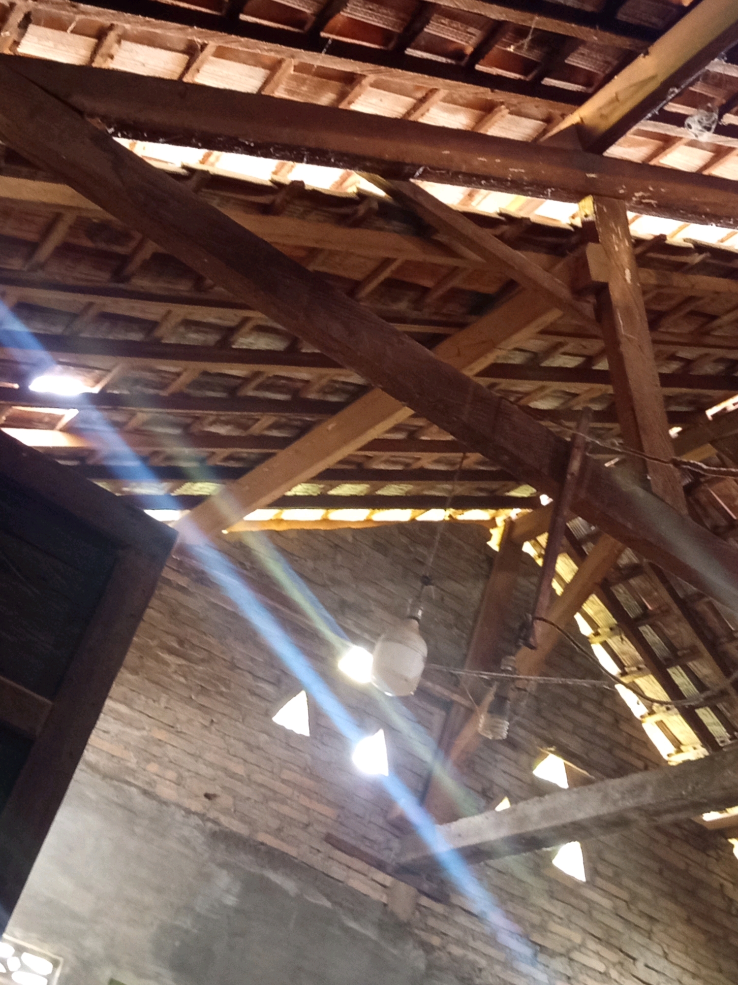 The image depicts the interior of a roof structure, likely an old building, in Karang Sari, Indonesia. The roof is constructed with wooden beams and covered with terracotta tiles, with some areas showing gaps that allow sunlight to penetrate. Two light bulbs are visible, hanging from wires. The walls are made of brick, and some parts are covered with cement plaster. Rainbow-like light flares are present, caused by the sunlight interacting with the camera lens. The overall impression is of a rustic and possibly neglected structure.