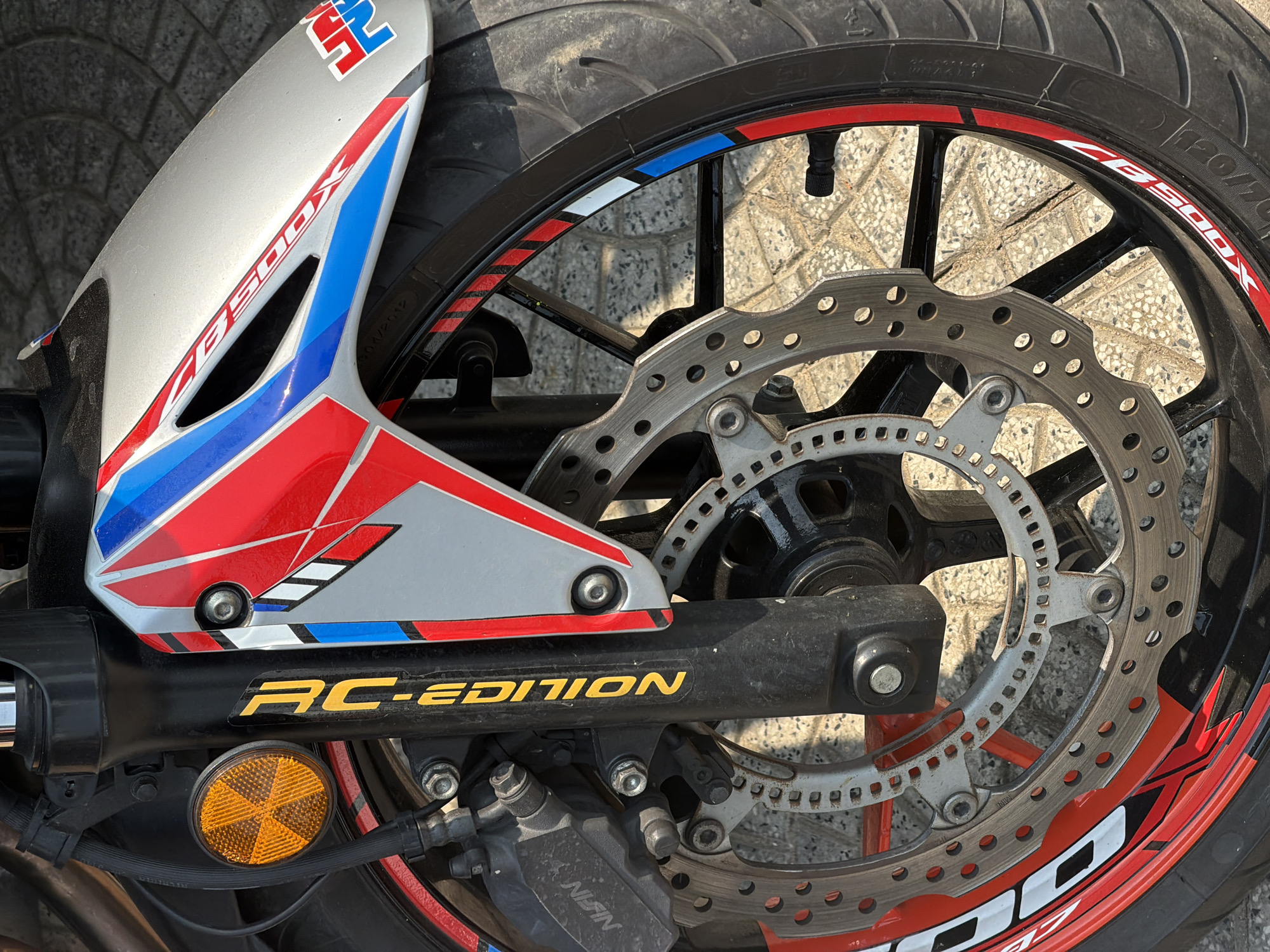 The image shows the rear wheel and swingarm of a Honda CB500X RC Edition motorcycle. The motorcycle is parked on a paved surface. Details visible include the rear tire, the brake discs, the "RC-EDITION" branding on the swingarm, and the orange reflector. Part of the rear mudguard, painted with red, white, and blue livery, is also visible. The motorcycle appears to be in Đà Nẵng, Vietnam based on common motorcycle models and the typical urban paving stones found in that area.