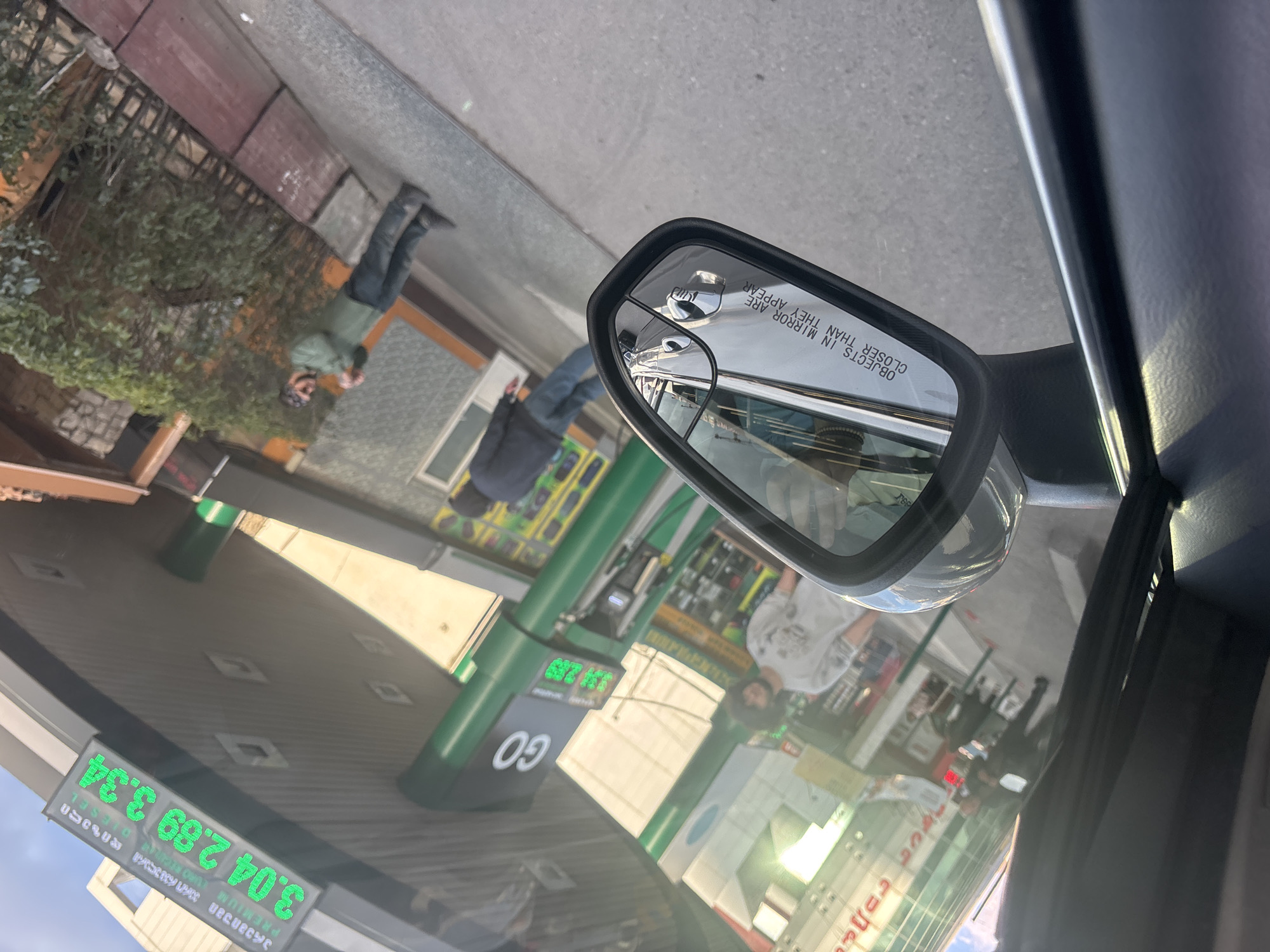 This image, taken from inside a car, shows a Go gas station reflected in the passenger side mirror. The gas prices are displayed as 3.04, 2.89, and 3.34. A person is visible outside the car, and a second person is reflected in the mirror looking down at their phone in front of a building. The surrounding environment suggests an urban area. Based on the gas station and the urban environment, it is possible this image was taken in Tbilisi, Georgia.