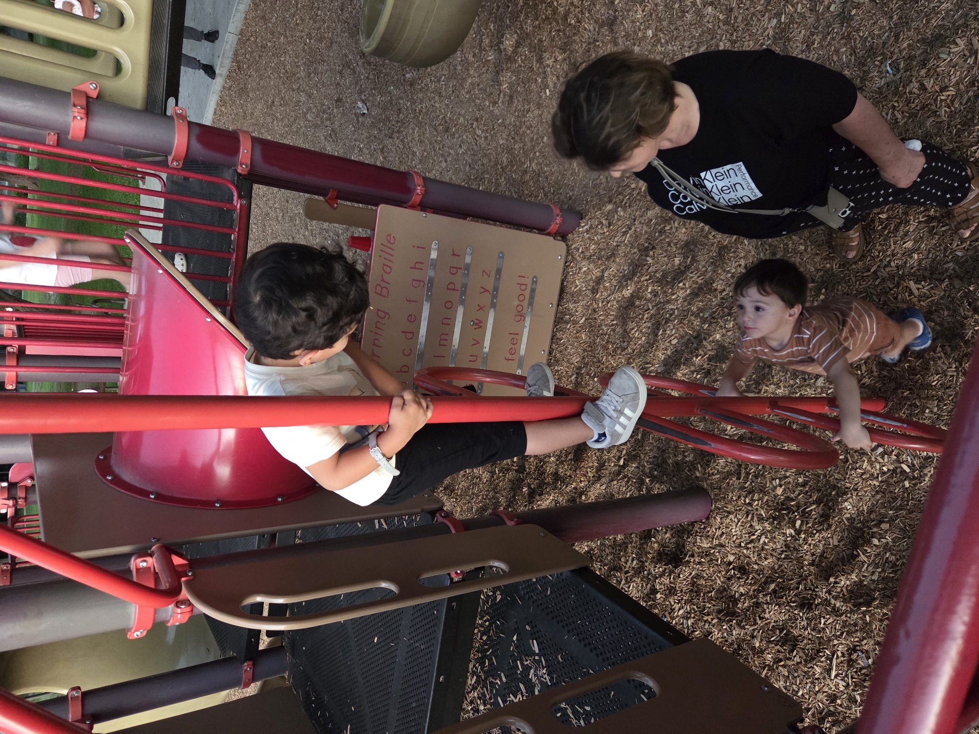 In Glendale, Arizona, a playground scene unfolds with three individuals. A young boy, wearing a striped t-shirt and blue shoes, stands looking upwards. To his right, an adult woman in a black shirt with a white graphic and patterned pants observes him. In the background, a slightly older boy in a white t-shirt and black pants is climbing a red play structure. A beige sign with braille characters and the words "feel good!" is attached to the structure. The ground is covered in wood chips, typical of a playground environment.