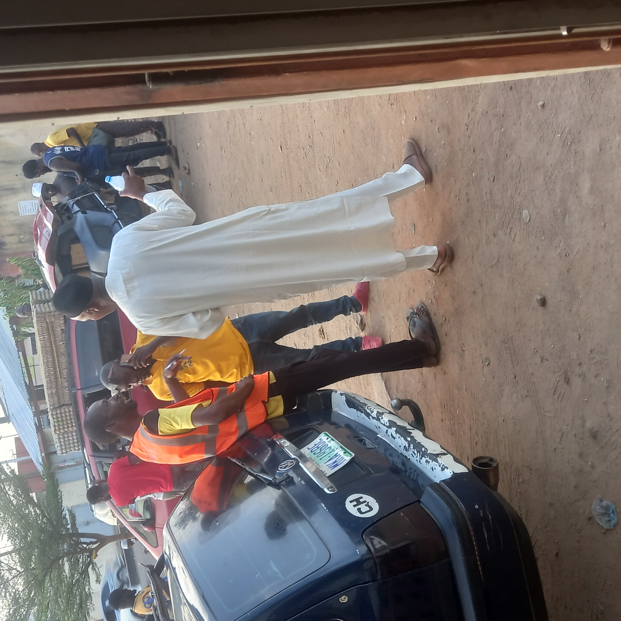 This image depicts an outdoor scene in Dadi Riba, Nigeria, likely during daytime under clear weather. A group of men are gathered on a dusty, unpaved ground surface, which appears to be reddish-brown earth.
In the foreground and midground, a man wearing a long white traditional robe (agbada) and a dark cap stands, holding a clear plastic water bottle. He faces towards other men in the group. To his right and slightly behind him, another man in a yellow t-shirt, blue jeans, and pink shoes is visible. Further back, another man in a yellow t-shirt and dark trousers, wearing brown sandals, stands.
To the left, a man in an orange reflective safety vest over a yellow t-shirt is leaning on the hood of a dark blue car. This car, possibly an older model, features a license plate clearly displaying "WWK-J36BE" and a white circular sticker with "CH" on its front. The car shows some wear, with paint appearing to peel on the edges of its top.
Further in the background, more individuals are partially visible, including a man in a blue t-shirt and dark trousers, and another in a red t-shirt. Portions of other vehicles, including a red car and what might be a motorcycle or tricycle, are also seen. A building structure and some green vegetation are visible on the far left. The individuals appear to be engaged in casual interaction or simply standing together, with neutral expressions. The overall impression is a social gathering in a public area.
