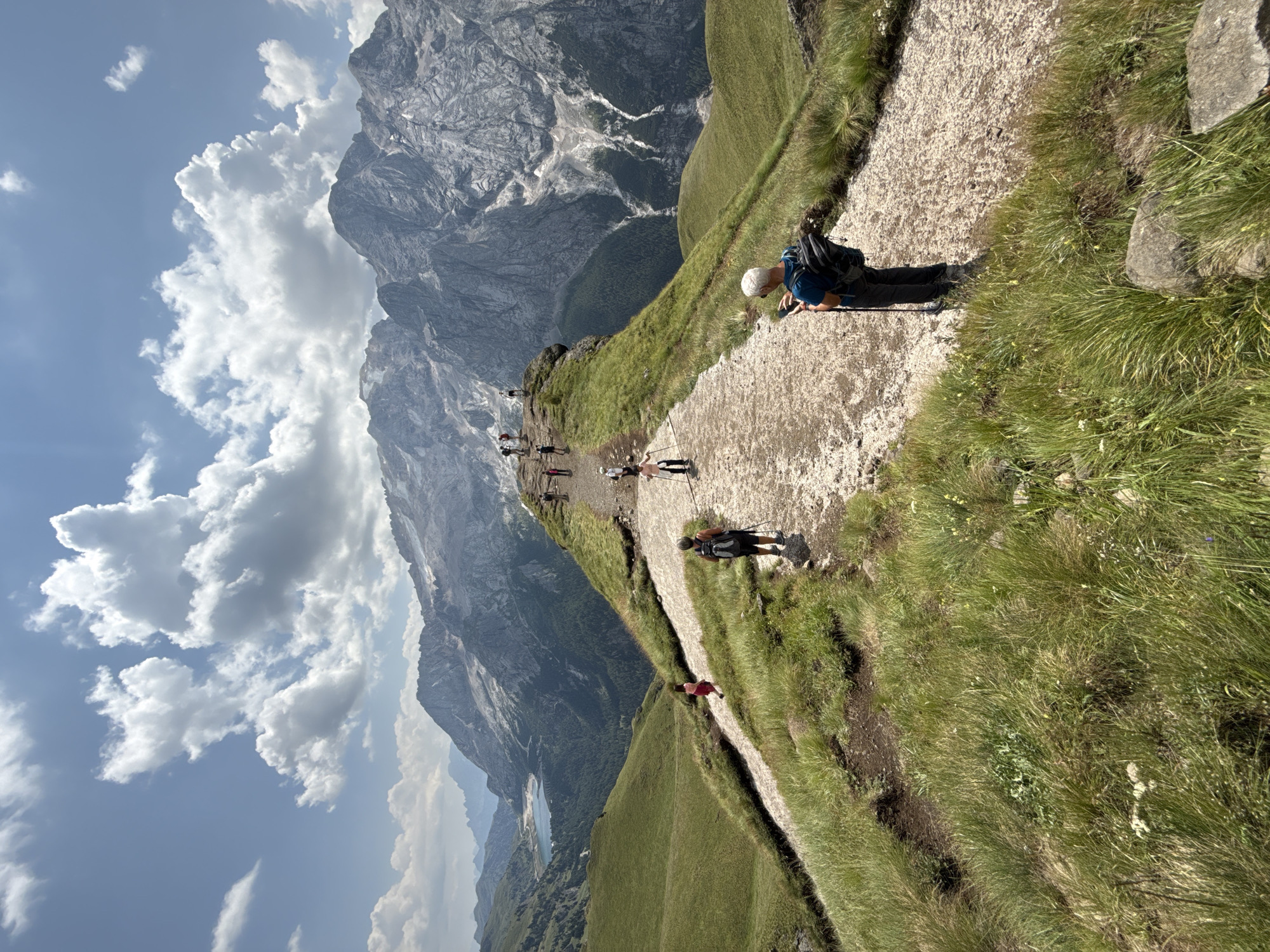 The image depicts a scenic mountain trail in the Dolomites, near Canazei, Italy. A group of hikers are visible on a gravel path that winds along a grassy ridge. In the foreground, a person with a backpack and walking sticks is looking at their phone. Further along the path, several other hikers are spaced out, some walking and some standing. In the background, a picturesque lake is nestled amongst lush green hills, and towering, rugged mountains with snow-capped peaks dominate the horizon under a partly cloudy blue sky. The environment is characterized by steep, verdant slopes and dramatic mountain formations, typical of the Alpine region.