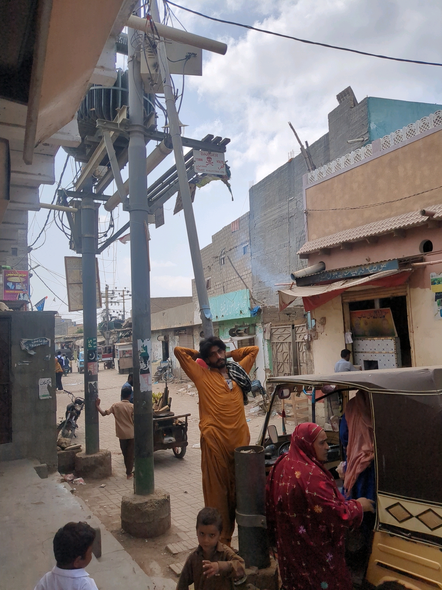 A street scene in Karachi Division, Pakistan, with a central focus on a utility pole featuring a large transformer and numerous electrical wires. A man in an orange outfit stands with his back to the camera, looking up at the sky. Several other individuals are scattered throughout the scene, including a woman in a red hijab entering an auto-rickshaw, a child in a brown shirt, and a child in a white shirt in the foreground. Various buildings, some with signs in Urdu, line the street, along with parked motorcycles and a pushcart. The overall impression is of a busy, somewhat cluttered urban environment under a cloudy sky.