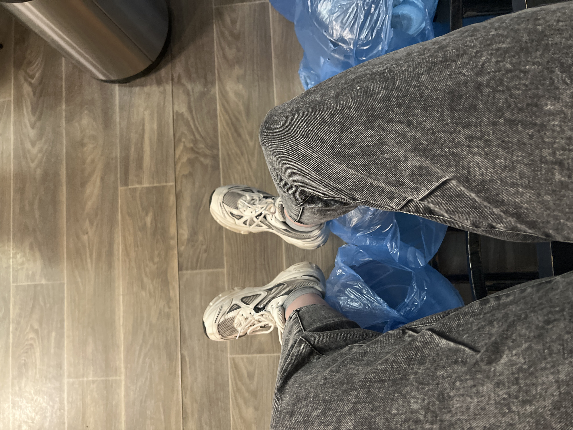 The image shows a person's legs and feet. The person is wearing gray jeans and white sneakers. The person is sitting on a chair with a blue plastic bag underneath. A stainless steel trash can is to the left. The floor is made of gray wood-look tiles. There is no evidence to suggest this photo was taken in Khimki, Russia.