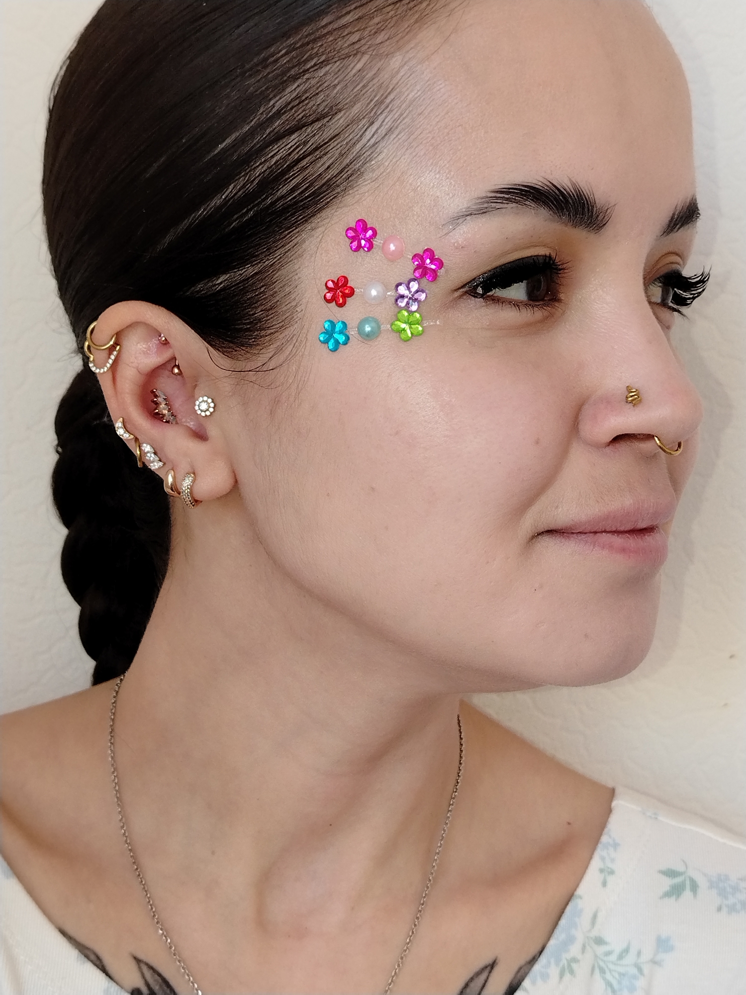 A close-up, portrait-style image features a woman with dark hair styled back from her face. The image is well-lit, showcasing the woman's fair skin tone.
The woman has a noticeable amount of facial jewelry. This includes colorful flower-shaped jewels and iridescent beads placed on her forehead near her temple, and multiple gold-colored piercings in her left ear.
She also has two nose piercings, both adorned with gold jewelry. She is wearing eyeliner and false eyelashes.
A silver chain necklace is visible on her neck. A faint tattoo is also visible below her neck.
The background is a plain, off-white wall, causing the subject to be in the foreground. There is no indication of the location being in the city of Noviy, Russia.