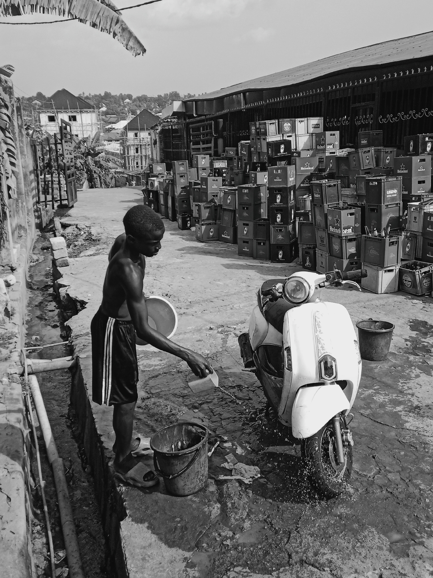 A black and white image taken outdoors in Onitsha, Nigeria, depicts a shirtless, dark-skinned young man in athletic shorts washing a white scooter. The man stands near a narrow ditch, using a small container to pour water from a bucket onto the front tire of the scooter. Adjacent to the scooter are stacks of beer crates, many bearing labels such as "Heineken," "Gulder," "STAR," and "Legend Extra Stout." In the background, buildings and structures are visible, hinting at the urban setting. The immediate surroundings of the man and scooter show signs of mud and water, suggesting recent or ongoing cleaning activities.