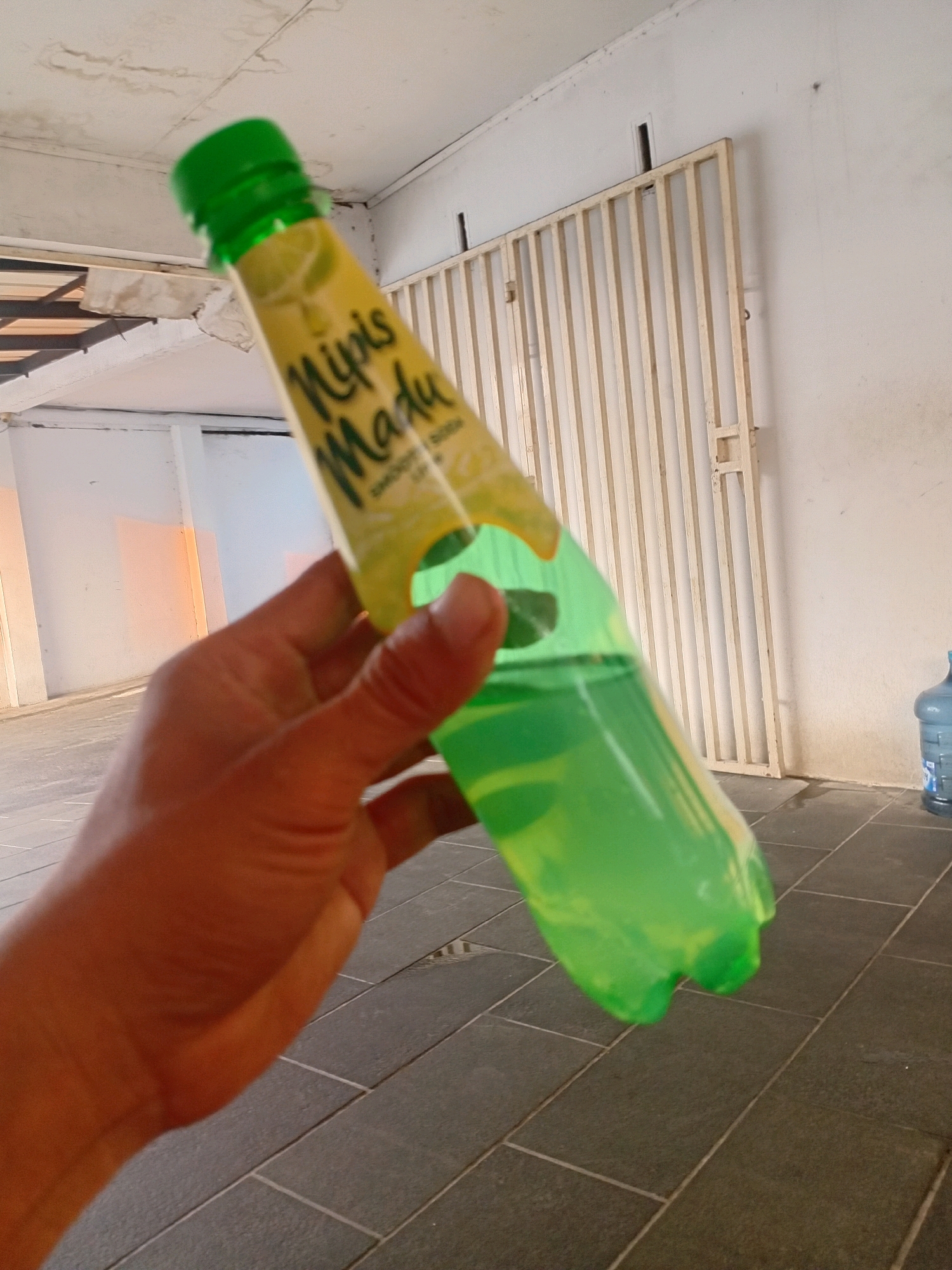 A person's hand is holding a green plastic bottle filled with a light green liquid, with a yellow label. The bottle appears to be a beverage. The background consists of a white wall with a beige metal grate. The floor is tiled with dark gray square tiles. A large blue water jug is visible in the far right background. The scene suggests an outdoor or semi-outdoor urban environment. The location is the city of Tangerang, Indonesia.