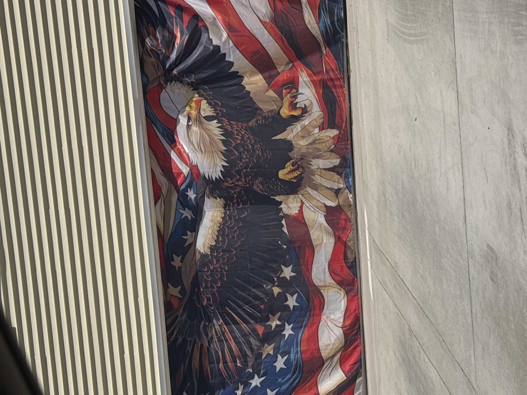 This media file shows a large banner featuring a bald eagle in flight against the backdrop of the American flag. The eagle, depicted with detailed feathers and sharp talons, appears to be soaring upwards. The American flag is stylized, with its red and white stripes and blue field with stars flowing and wrapping around the eagle.
The banner is hanging vertically and is partially obscured by a fluted building facade on the left and a rough, textured concrete wall on the right. The lighting suggests it is daytime, with sunlight casting shadows and highlights on the building materials. The scene appears to be outdoors, possibly on the side of a building.
There is no visible text in the image. The overall impression is one of patriotism and strength, with the eagle symbolizing freedom and the flag representing the United States. The setting seems to be a public space in Citrus Heights, USA, given the location context. The weather appears to be clear.