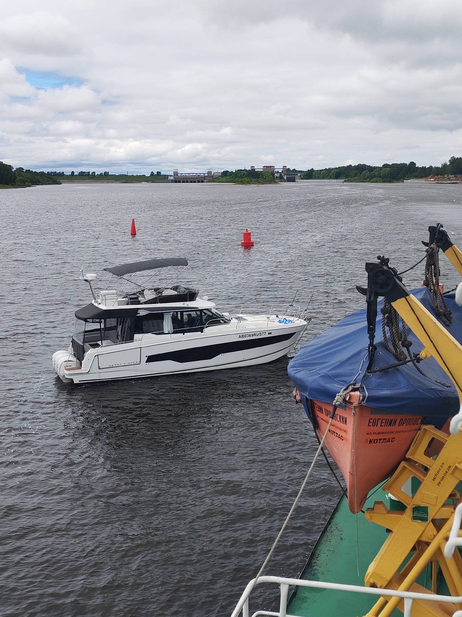 The image depicts a river scene. In the foreground, there is a section of a vessel with a lifeboat covered by a blue tarp and secured by yellow davits. The lifeboat is painted orange and has lettering in Cyrillic script. A white modern yacht with a black hardtop is positioned in the midground, featuring a black stripe along its hull and the registration number "AB0369RUS77." Two red navigational buoys are visible in the water between the two boats. In the background, a river lock or dam structure is situated on the horizon line, with green riverbanks and foliage flanking the water's edge. The sky is overcast with gray and white clouds. The specific location in the city of Unknown, Unknown, cannot be determined.