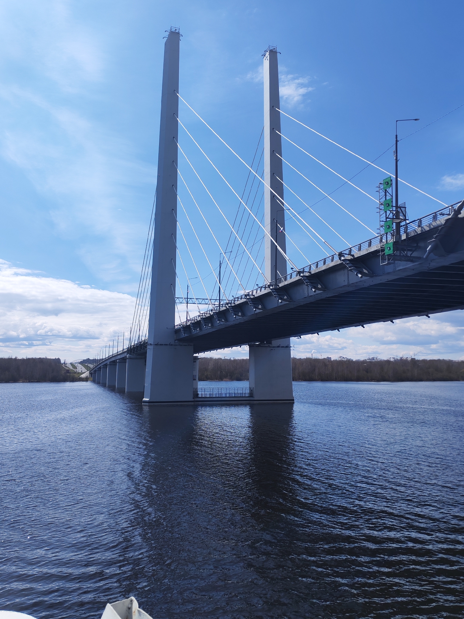The image displays a modern cable-stayed bridge spanning a wide, dark blue river under a bright, partly cloudy sky. Two imposing, light grey concrete pylons, rectangular in cross-section, rise from the river, anchoring numerous white tension cables that fan out to support the grey bridge deck. The bridge features streetlights along its length and, on the right side, green lattice-like structures are visible on the side of the deck near one of the pylons.
The river's surface is rippled, reflecting the sky and the bridge's structures, with deeper shadows closer to the bridge's foundations. The far riverbank is lined with a dense growth of trees, which appear bare or sparsely foliated, suggesting either early spring or late autumn. A road or highway can be seen ascending into the distance on the far side of the river. The sky is a vibrant blue with scattered white cumulus clouds, indicating clear and fair weather conditions during what appears to be daytime. No people are visible. This scene is located in Cherepovets, Russia. There is no visible text.