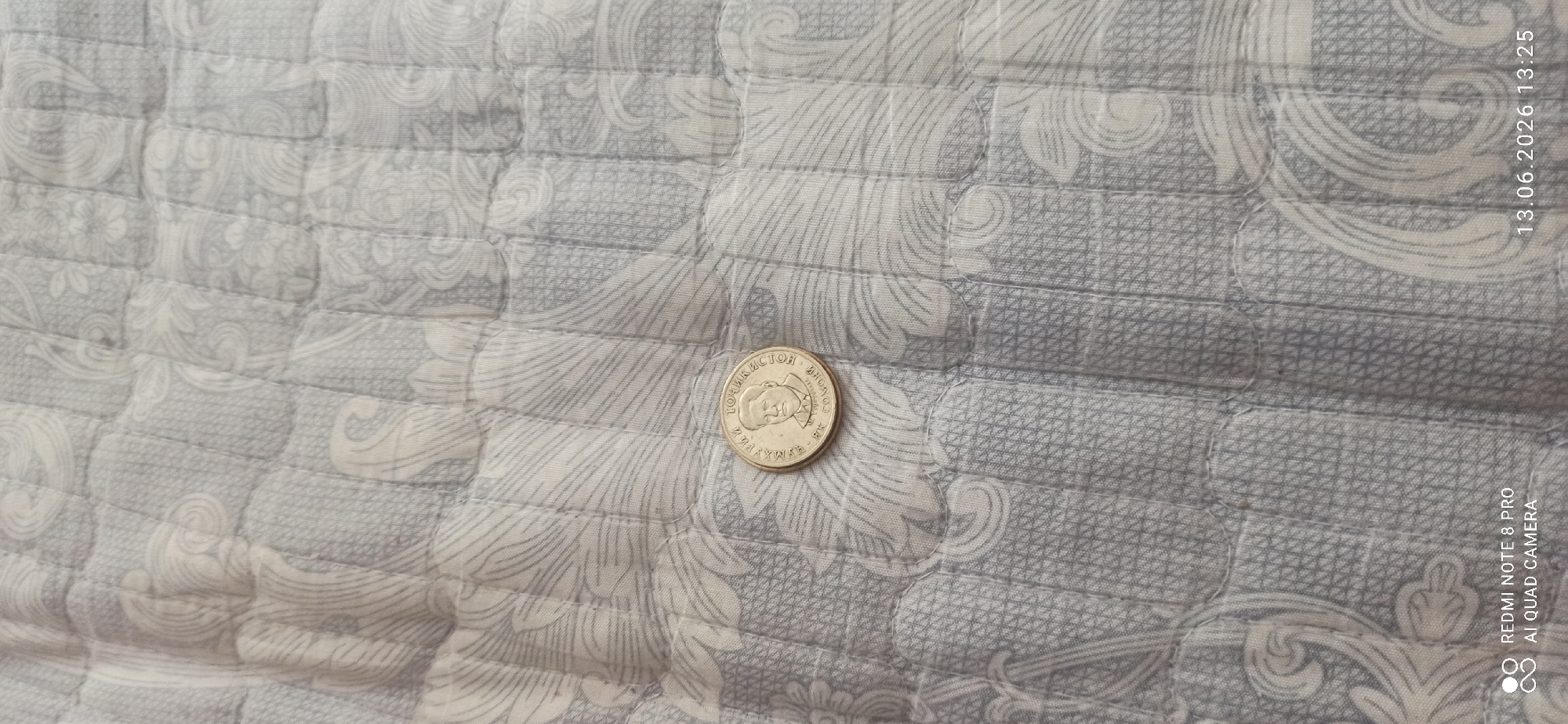 An overhead medium shot captures a single coin resting on a patterned quilted surface. The coin is a light metallic color and features a profile portrait of a man on its obverse. Text encircles the portrait. The quilted surface is predominantly light gray with a white swirling floral pattern. In the lower left corner, a watermark reads "REDMI NOTE 8 PRO AI QUAD CAMERA". In the lower right corner, a timestamp reads "13.06.2026 13:25". The scene appears to be indoors, on a bed or sofa.