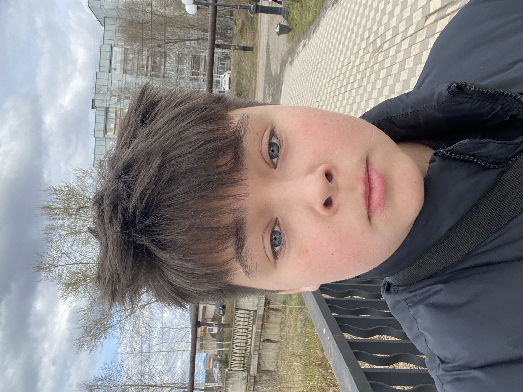 A young person, likely a child or pre-teen, is featured in a close-up selfie, looking directly at the camera with a neutral expression. They have bright blue eyes, fair skin with some pinkness on the cheeks and lips, and short, dark brown hair that falls over their forehead. They are wearing a dark blue or black puffy jacket with a visible strap across the shoulder, possibly from a bag.
The setting is outdoors on an overcast day, with a grey, cloudy sky dominating parts of the background. Bare trees with slender branches are scattered throughout, suggesting either early spring or late autumn. In the mid-ground, there are multi-story buildings, some with reflective glass windows, indicating an urban or semi-urban environment. A paved path with a distinct checkerboard or diamond pattern is visible on the right, alongside a dirt or gravel path on the left. Two lampposts or streetlights stand in the upper right background. A dark, wavy metal railing or fence is present in the lower right foreground. A white vehicle is partially visible in the distant left background. The overall scene suggests a public outdoor space in Votkinsk, Russia. There is no visible text in the image. The original image is rotated 90 degrees clockwise.
