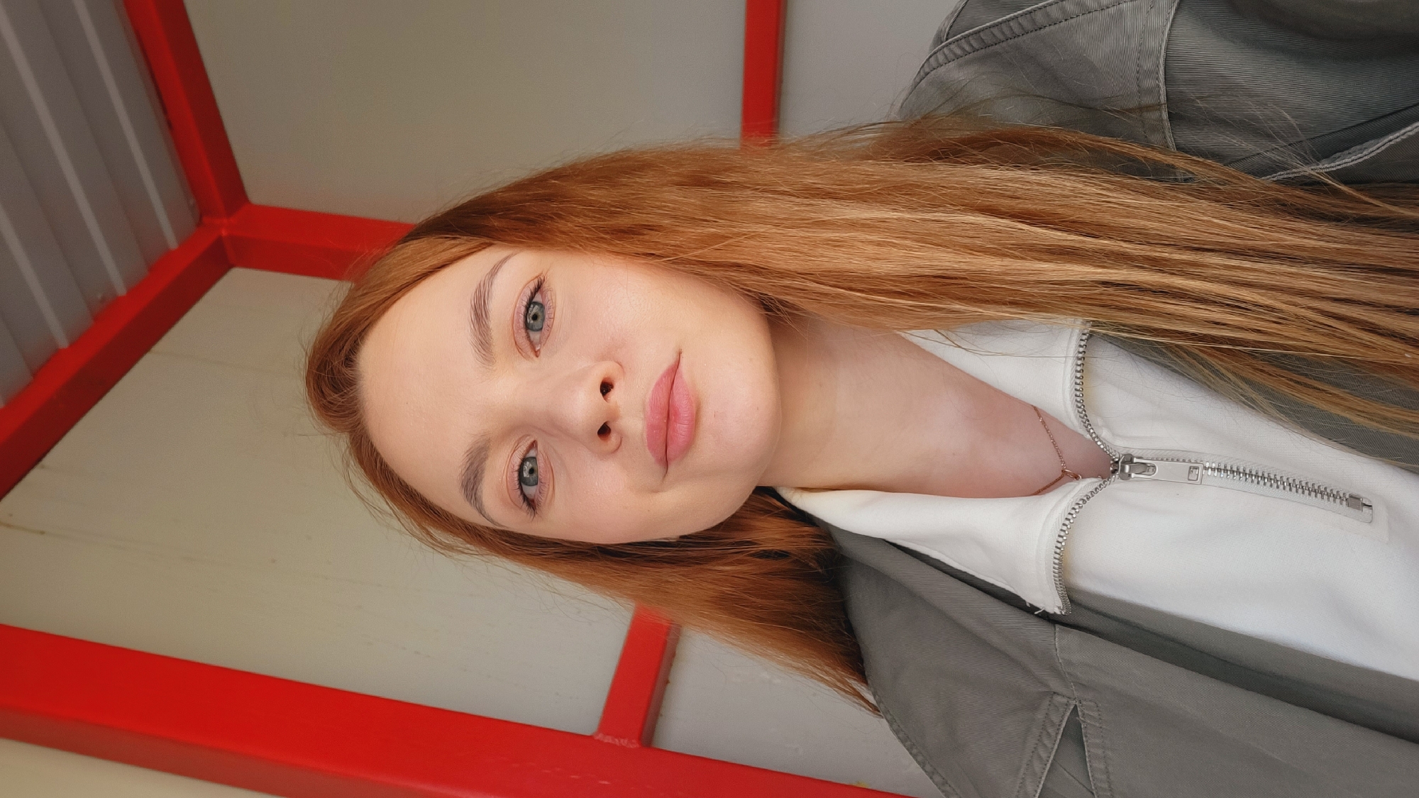 A woman with long, red hair is photographed from below. She is wearing a white shirt and a green jacket. Behind her is a red metal structure. The location is indicated as Chaikovsky, Russia, but no elements visible in the image definitively confirm this.