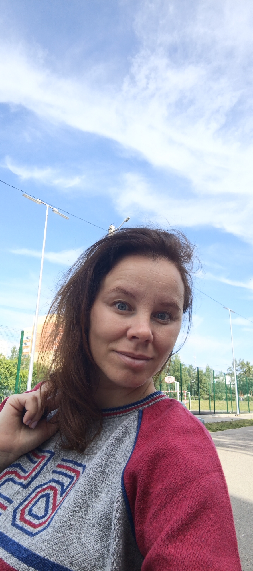 A woman with brown hair and blue eyes is taking a selfie outdoors. She is wearing a gray sweater with red sleeves and a blue and red design on the front. In the background, there is a blue sky with clouds, streetlights, a fence, trees, and a basketball hoop. The location is the city of Chaikovsky, Russia. The woman is looking at the camera with a slight smile. Her left hand is touching her hair.