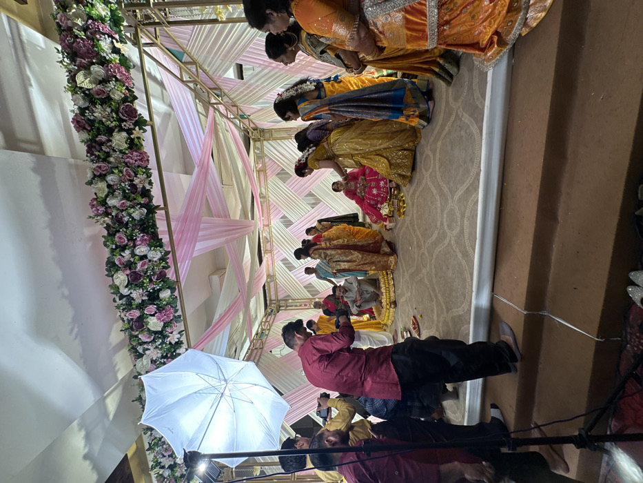 A wedding ceremony is underway beneath a floral-decorated arch with pink and white fabric. Several people in traditional Indian clothing are bowing. A photographer in a maroon shirt is taking pictures, his flash umbrella is in the foreground. Additional individuals are standing behind him. The scene is indoors, with an off-white patterned carpet.