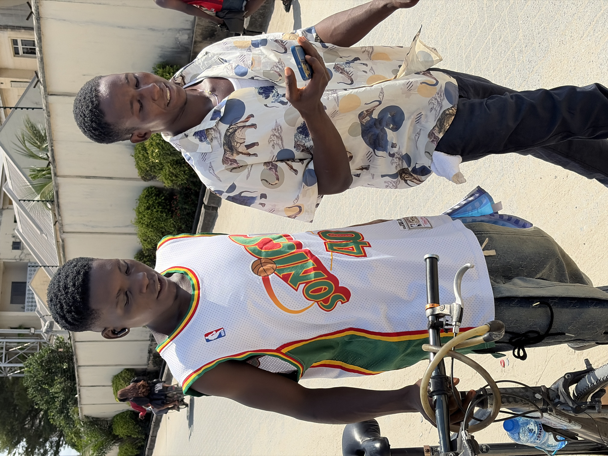 The image shows two individuals in what appears to be a city setting, likely Igboefon, Nigeria. One person is wearing a white, green, and red basketball jersey with the word "SONICS" displayed. The other individual is wearing a white shirt with animal print patterns and dark pants. The person in the animal print shirt is looking at and holding a small blue object. A bicycle is present and there are people in the background. There is light colored building. The ground is concrete or paved. The setting is outdoors, in daylight.