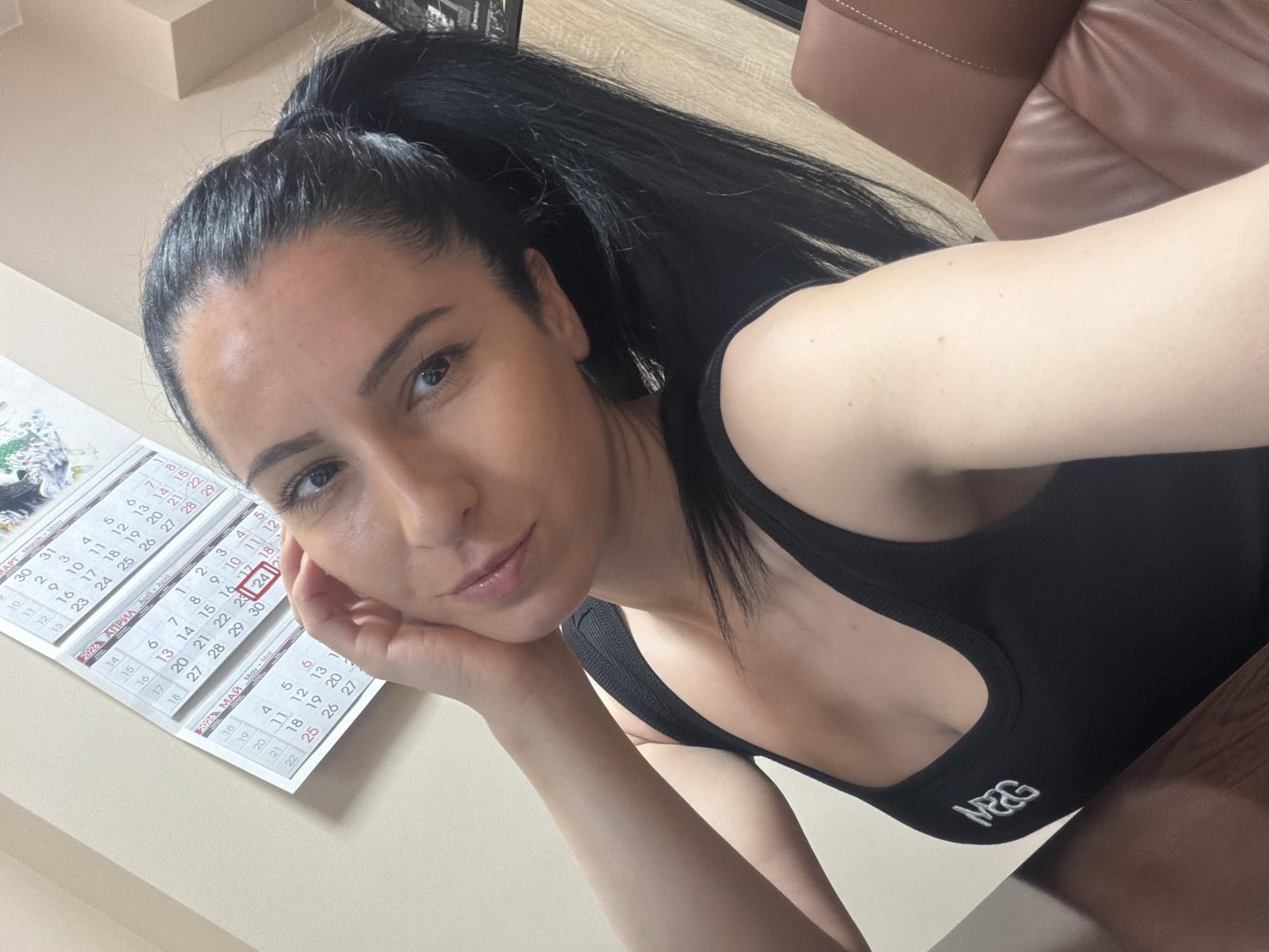 This media file features a woman taking a selfie in an indoor setting, likely an office or home in Pleven, Bulgaria. She is captured from the chest up, with dark hair pulled back into a high ponytail and a relaxed, neutral expression, resting her chin on her hand as she looks directly at the camera. She wears a black top or sports bra with a white "MRG" logo visible on the front.
In the background, to the left of the woman, a calendar is affixed to a light-colored wall. The calendar shows months in both Cyrillic and Latin script, including "МАРТ March", "АПРИЛ April", and "МАЙ May", for the year "2024". The date "24" in April is distinctly circled in red, potentially indicating the current date or a highlighted event. To the right, a warm brown leather armchair or sofa is partially visible, along with a wooden surface, suggesting a desk or table. The lighting appears natural and bright, indicating it is daytime. The Cyrillic text on the calendar is consistent with the provided location context of Bulgaria.
