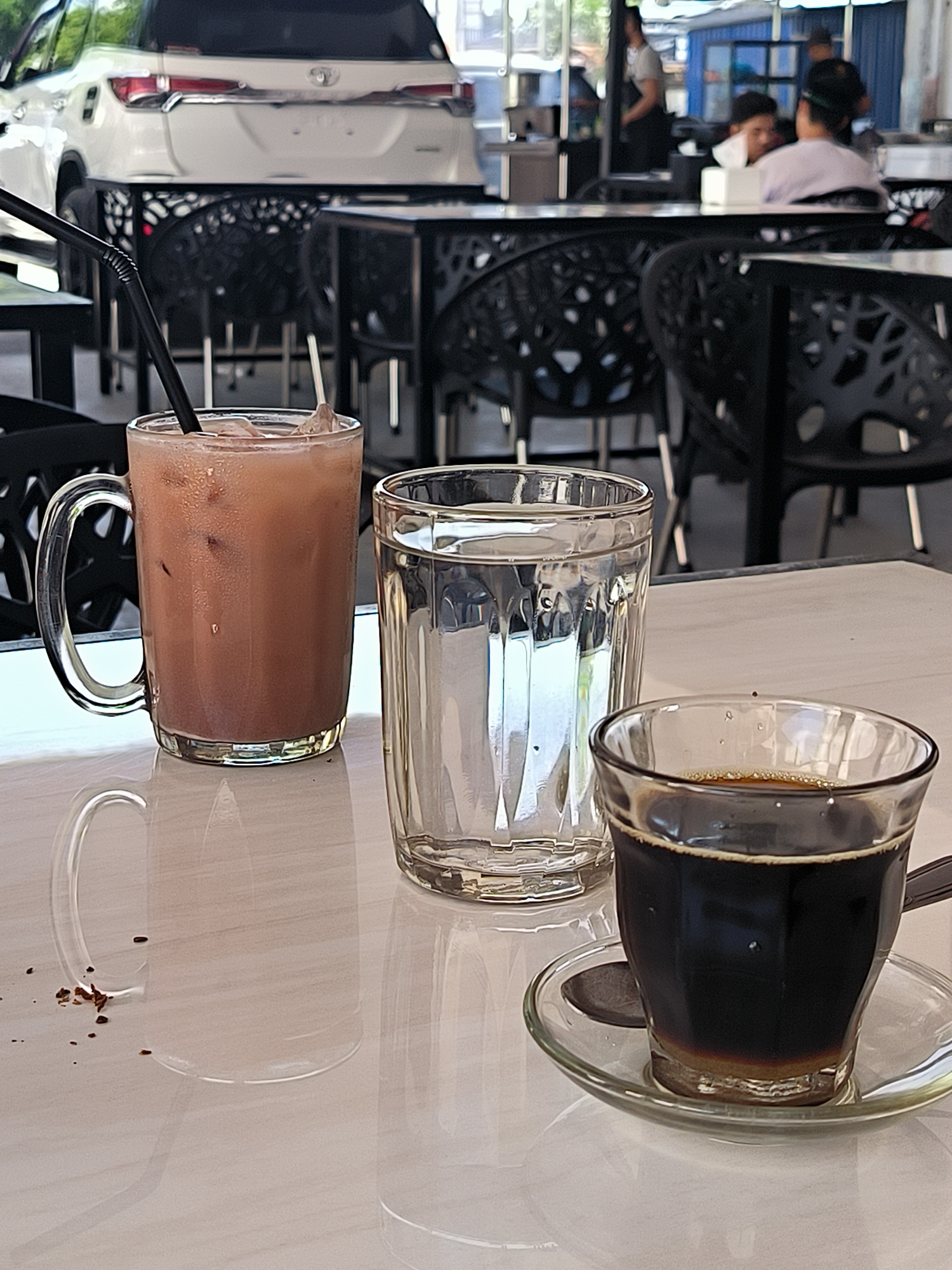 The image depicts a table setting in a cafe, possibly located in Lhokseumawe, Indonesia. On the table are three glasses: one with a brown beverage and a straw, another filled with water, and a small glass cup of black coffee with a saucer. Coffee grounds are scattered near the base of the first glass. In the background, people are sitting at other tables, with a white SUV visible. The scene suggests an outdoor dining area. The chairs and tables are made of dark-colored metal.
