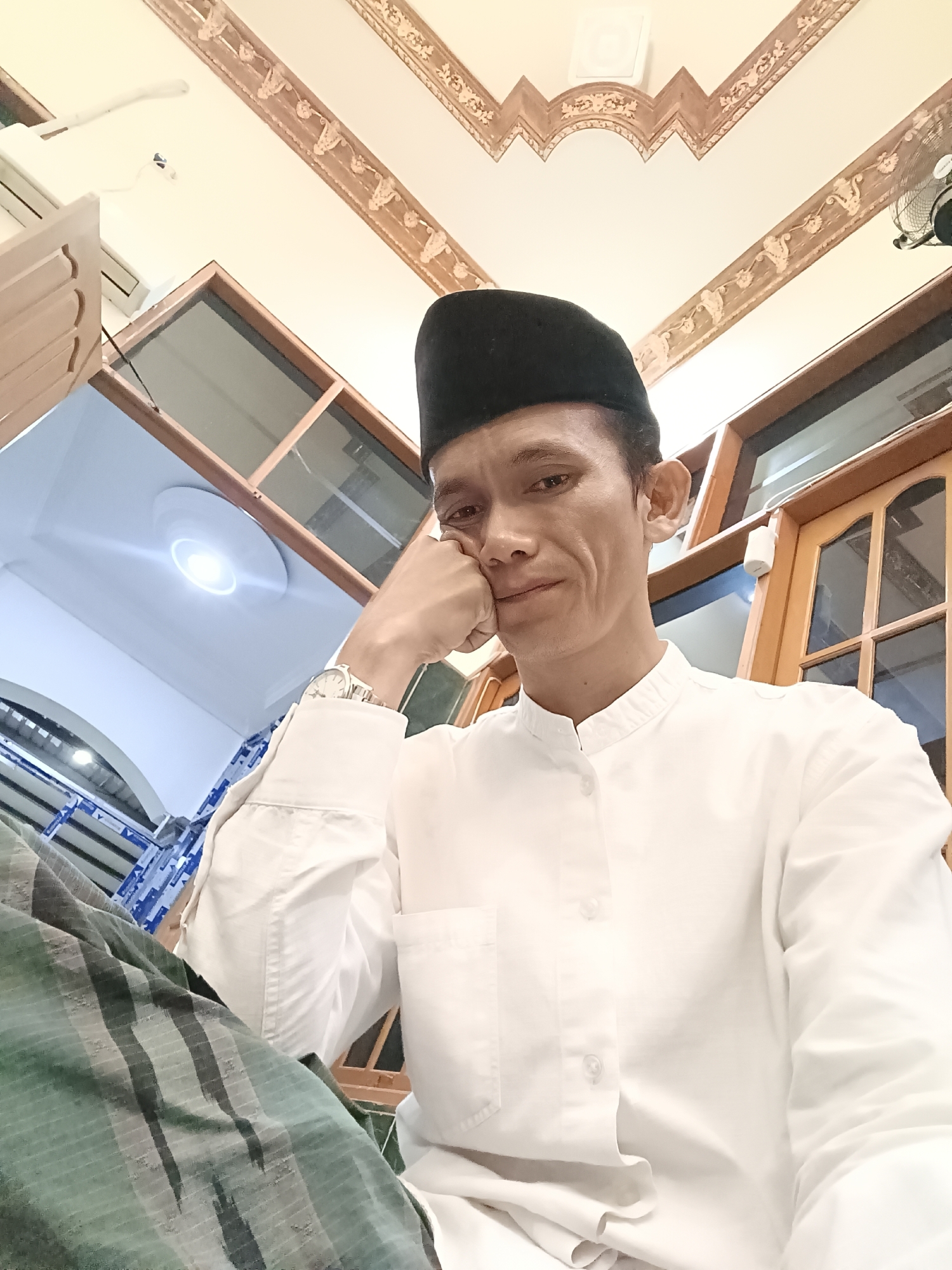 A man wearing a black peci and a white collared shirt is seated indoors, looking at the camera. He rests his right hand on his chin, with his elbow supported by his left hand which is hidden by his arm. A silver wristwatch is visible on his left wrist. He appears to be wearing a sarong with a green and dark gray striped pattern. The background features a ceiling with ornate wooden decorative molding. To the left, a light fixture is visible on the ceiling, and an air conditioning unit is mounted high on the wall. To the right, wooden framed windows and a ceiling fan are present. The environment suggests an indoor setting, possibly a prayer room or a common area within a building in Kediri City, Indonesia, given the presence of Islamic attire and typical Indonesian architectural elements.