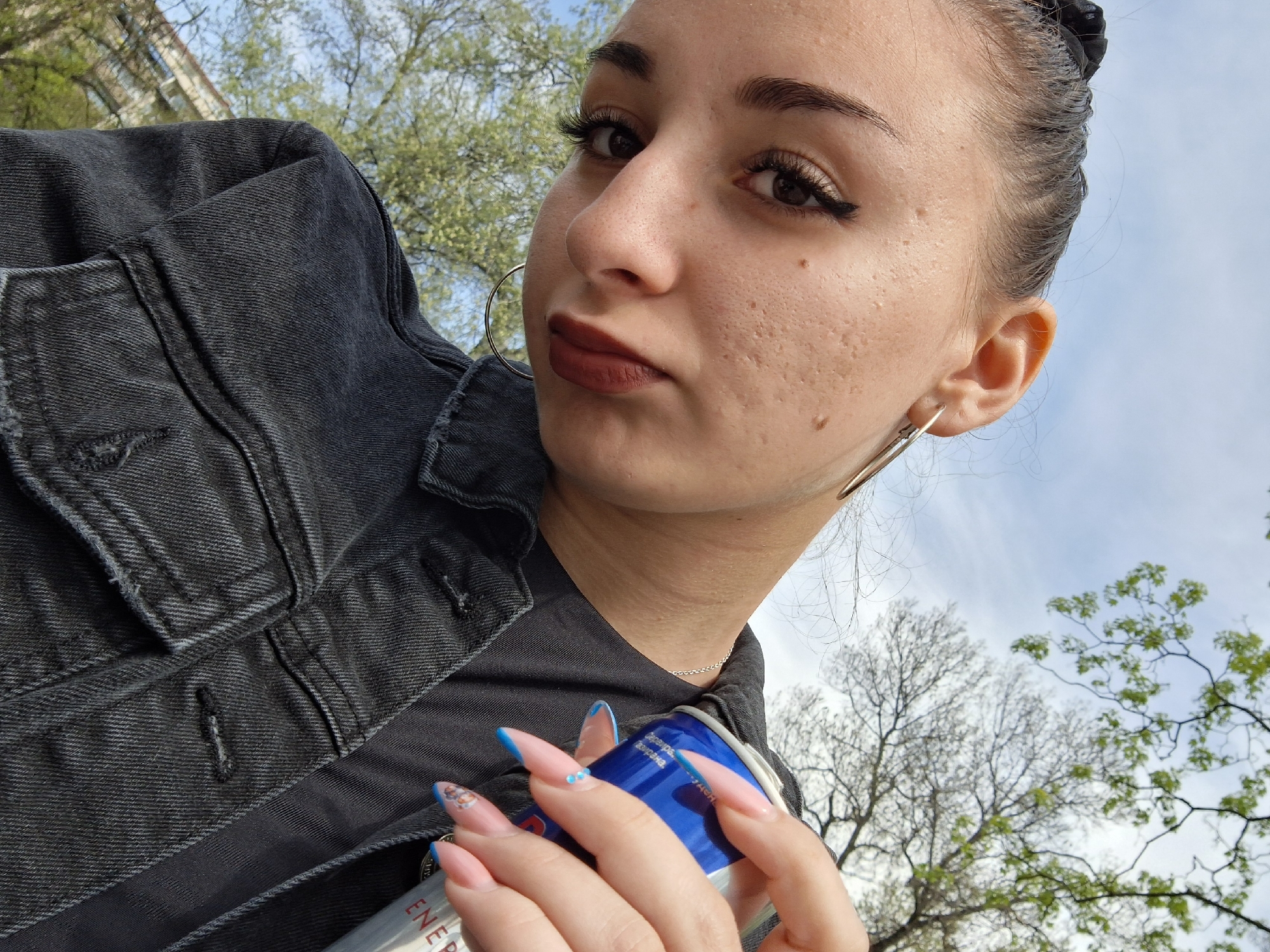 A woman is holding a blue and silver Red Bull energy drink can, while wearing a dark gray denim jacket and dark top. Her hair is pulled back. She is wearing silver hoop earrings and has long, almond-shaped nails with light pink polish and a blue tip. She has dark eyebrows, dark eyeliner, and dark lipstick. In the background is a blurry view of trees and a building, against a light blue sky. The location appears to be outdoors and could potentially be in Sofia, Bulgaria.