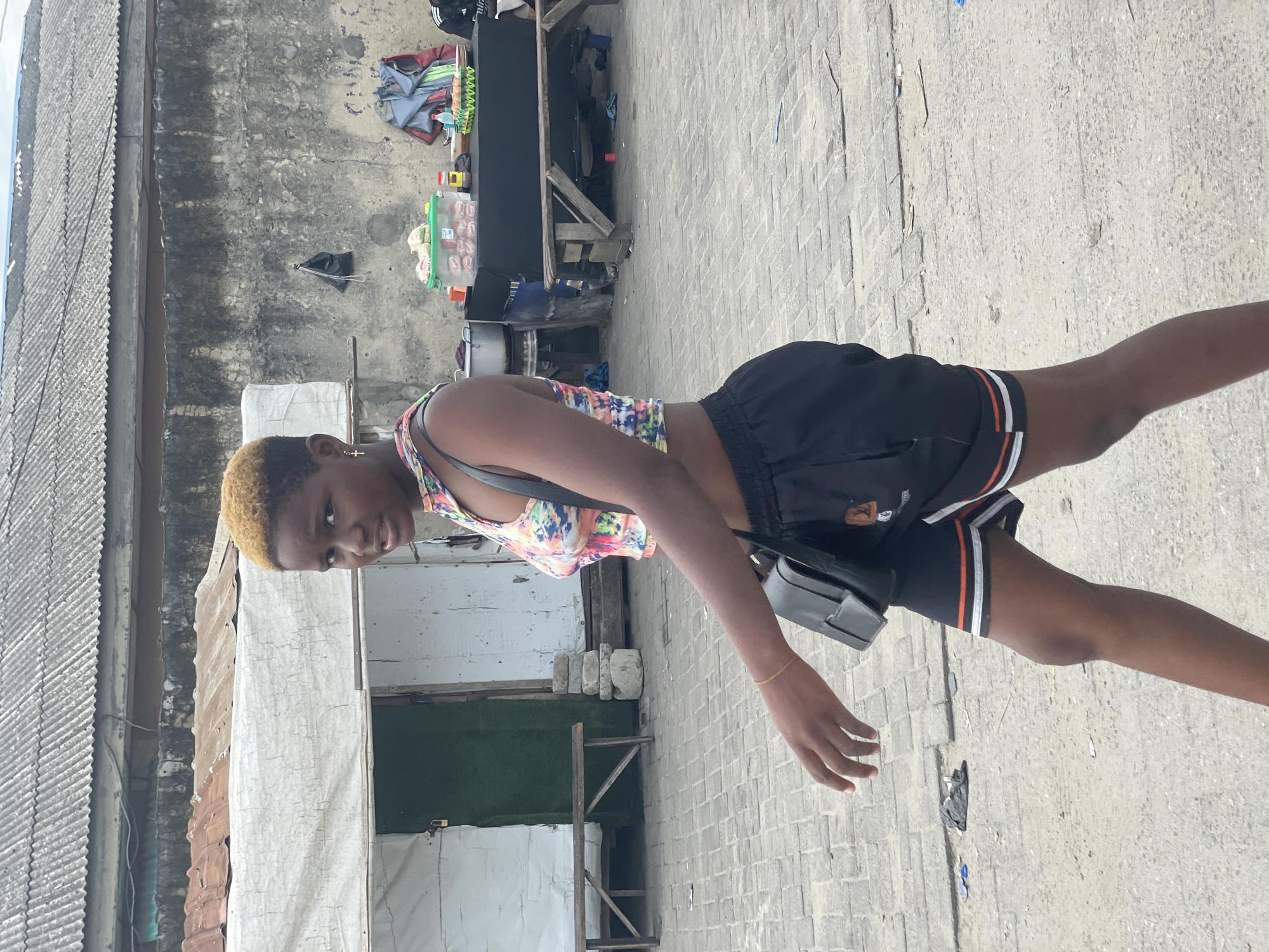 A full-body, portrait-oriented shot depicts a young person, likely female, standing on a paved street or pathway during bright daylight. She has short, light-colored (blonde or light brown) hair and is wearing a colorful, patterned sleeveless top with black shorts that feature white and orange stripes on the sides. A black shoulder bag is slung over her left shoulder, and a thin gold-colored bracelet adorns her right wrist. A small gold cross pendant is visible around her neck. She looks directly towards the camera with a neutral expression.
The background reveals a simple, urban or semi-urban setting. To the left, a building with a corrugated iron roof and wall is visible. To the right, a weathered concrete wall shows signs of discoloration and faint markings. Behind the subject, two makeshift wooden stalls or tables are set up. One is covered with black fabric, while the other displays various items, including packaged goods in clear plastic containers and a green basket filled with white packets, suggesting a street vendor's setup. A dark bag with a silver chain hangs on the concrete wall. The ground is made of concrete blocks and appears somewhat worn, with minor debris scattered about. The clear, sunny weather is evident from the bright lighting and distinct shadows. The scene, with its functional architecture and street vending, aligns with typical streetscapes found in Ado, Nigeria. A small, unidentifiable logo is on her shorts, and a faint "Emirates" logo can be spotted on an item in the far background.