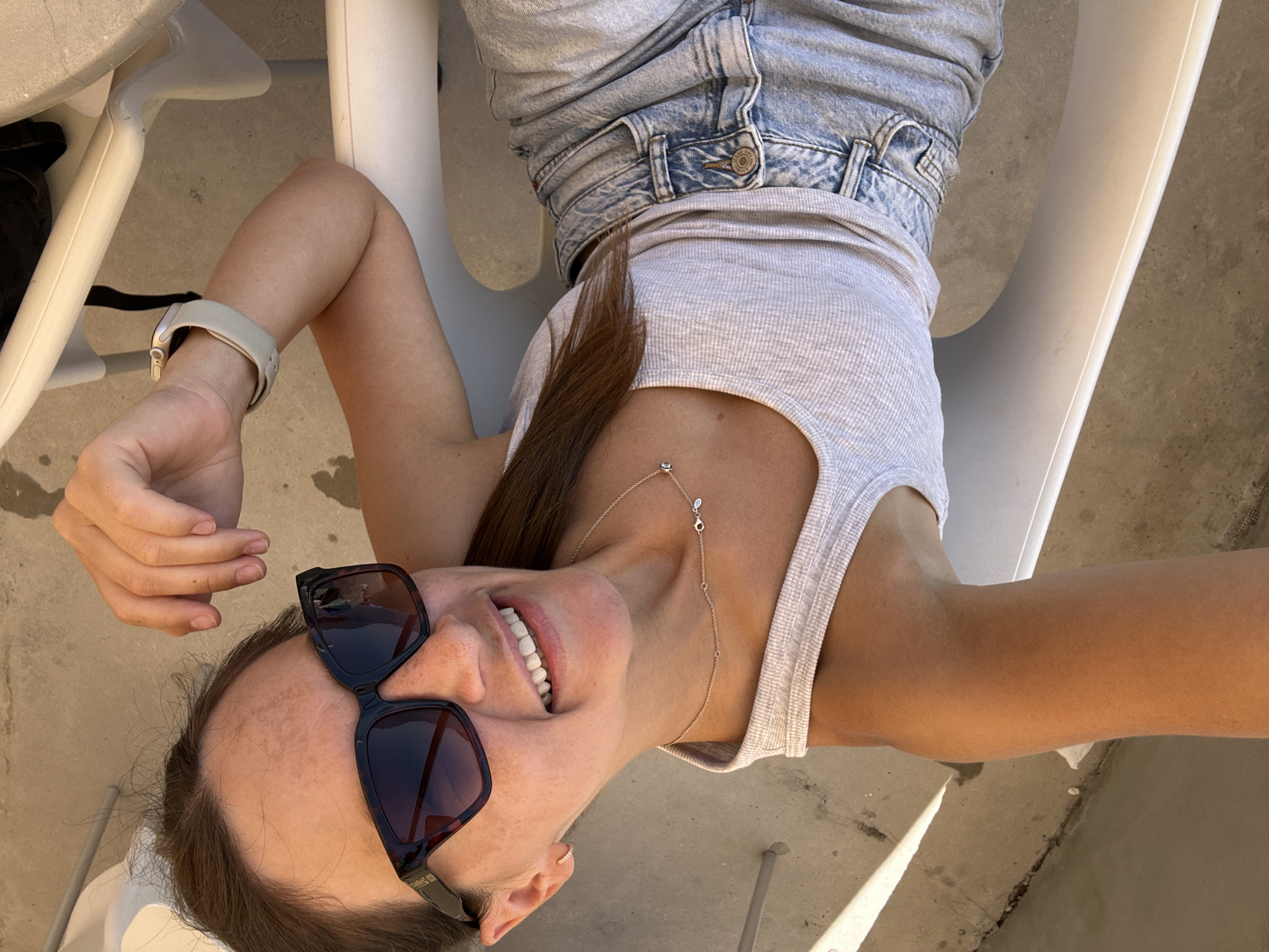 A young woman is lying on a white plastic lounge chair outdoors, with her head resting on her arms. She is wearing a grey ribbed tank top, light-wash denim shorts, and dark-rimmed sunglasses. A silver necklace with small charms is visible around her neck. A smartwatch with a light-colored band is on her left wrist. Her fingernails are unpainted.
The woman is smiling broadly and looking up towards the camera. Her eyes are open, and her teeth are visible. The lighting suggests it is daytime, likely sunny, as indicated by the strong shadows cast on the concrete ground. The setting appears to be a patio or poolside area. The concrete ground has some cracks and small pebbles.
The image is oriented sideways. No visible text is present in the image. The overall mood conveyed is relaxed and joyful, possibly indicating a vacation or leisure activity.