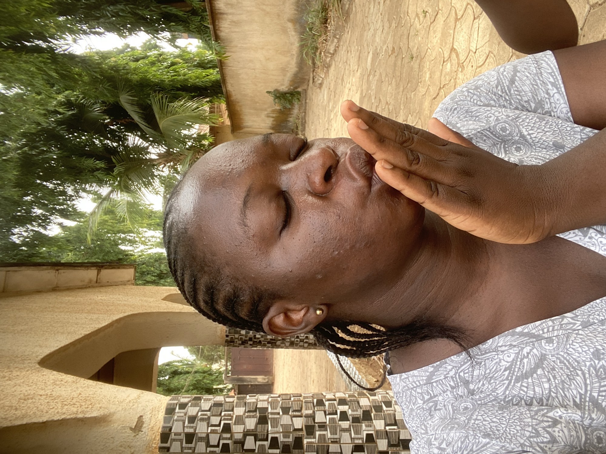 A woman with braided hair and closed eyes is positioned outdoors, with her hand raised near her face. She wears a patterned, light-colored garment. The background shows parts of a building with an archway and mosaic tiling, as well as trees and a pathway. The setting appears to be in Maigizo, Nigeria.