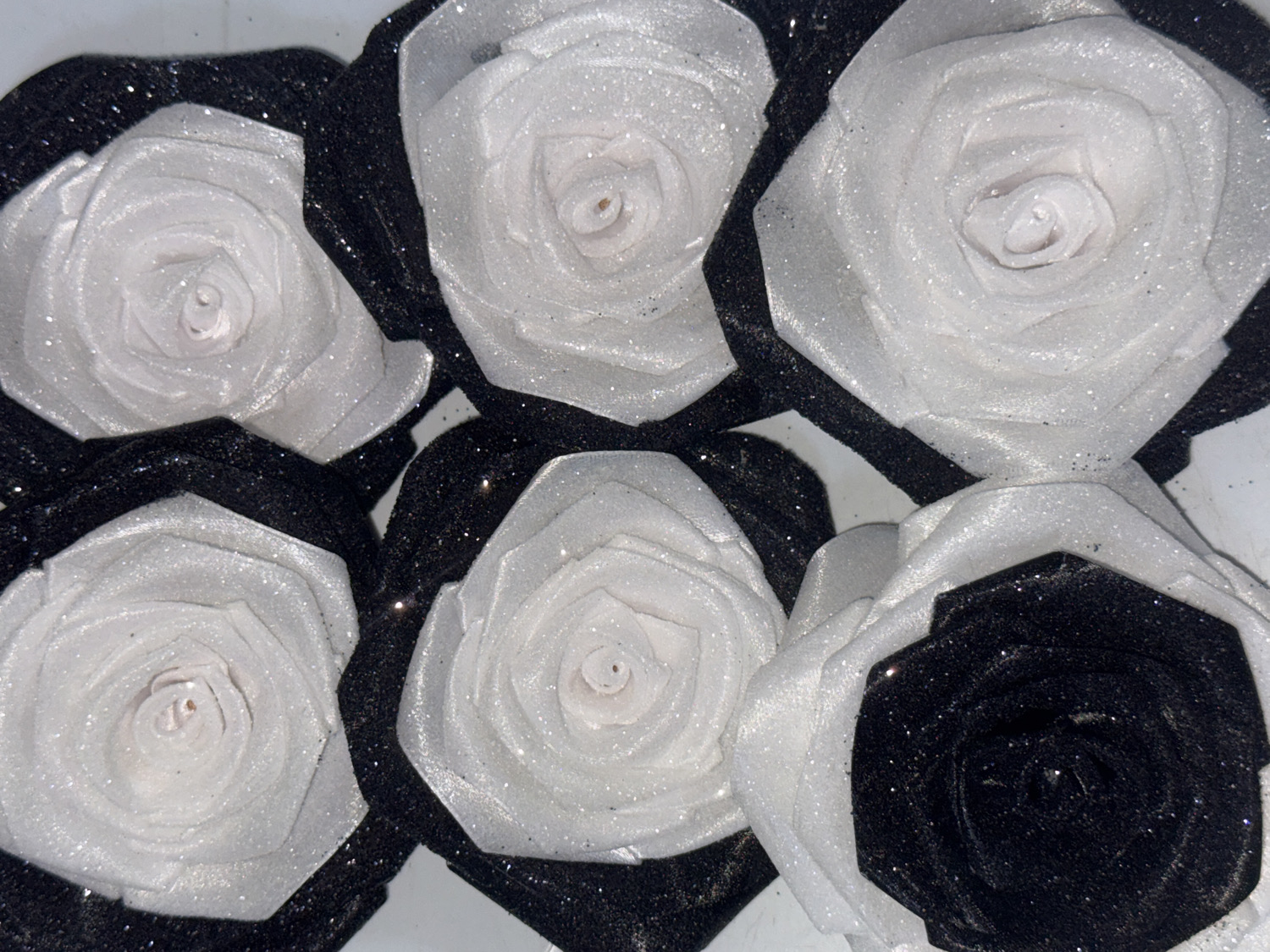 A close-up, overhead shot reveals an array of artificial roses, predominantly in a striking black and white color scheme, arranged closely together. The roses are crafted from fabric, likely satin or a similar material, and are generously dusted with silver glitter, giving them a shimmering appearance.
The arrangement features several white roses with distinct black outer petals, creating a checkerboard or alternating pattern. Each white rose displays intricate, layered petals that spiral inwards towards the center, where the core of the bloom is visible. The glitter adheres to both the white petals and the contrasting black outer layers, catching the light.
One prominent rose in the lower right portion of the frame is entirely black and heavily glittered, surrounded by white petals that serve as its outer layer. This single black rose stands out against the predominantly white-centered blooms. The texture of the glitter is clearly visible, adding depth and sparkle to the artificial flowers. The background is a soft, light color, possibly white, which helps the roses stand out. The image captures a decorative display, possibly for an event or craft, from Davao City, Philippines. There is no visible text.