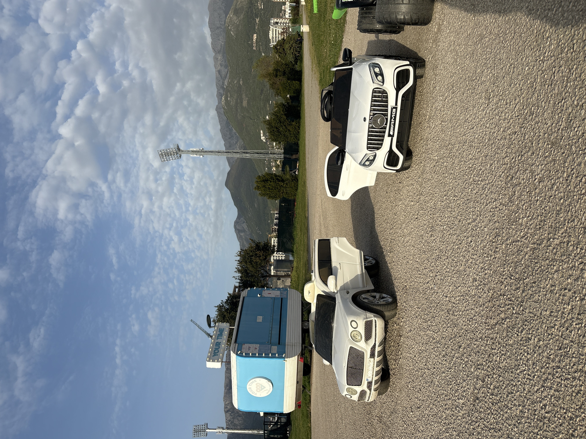 The image shows a paved area, possibly a parking lot or road, with two toy cars – a white Mercedes-AMG and a white Bentley – and a blue and white trailer labeled "YOUR TIME". Behind the vehicles, there's a green grassy area, trees, buildings, and mountains in the background. A tall metal light tower is visible on the left side of the image. The location is likely in the city of Bar, Montenegro, based on the mountainous terrain and architecture in the background.