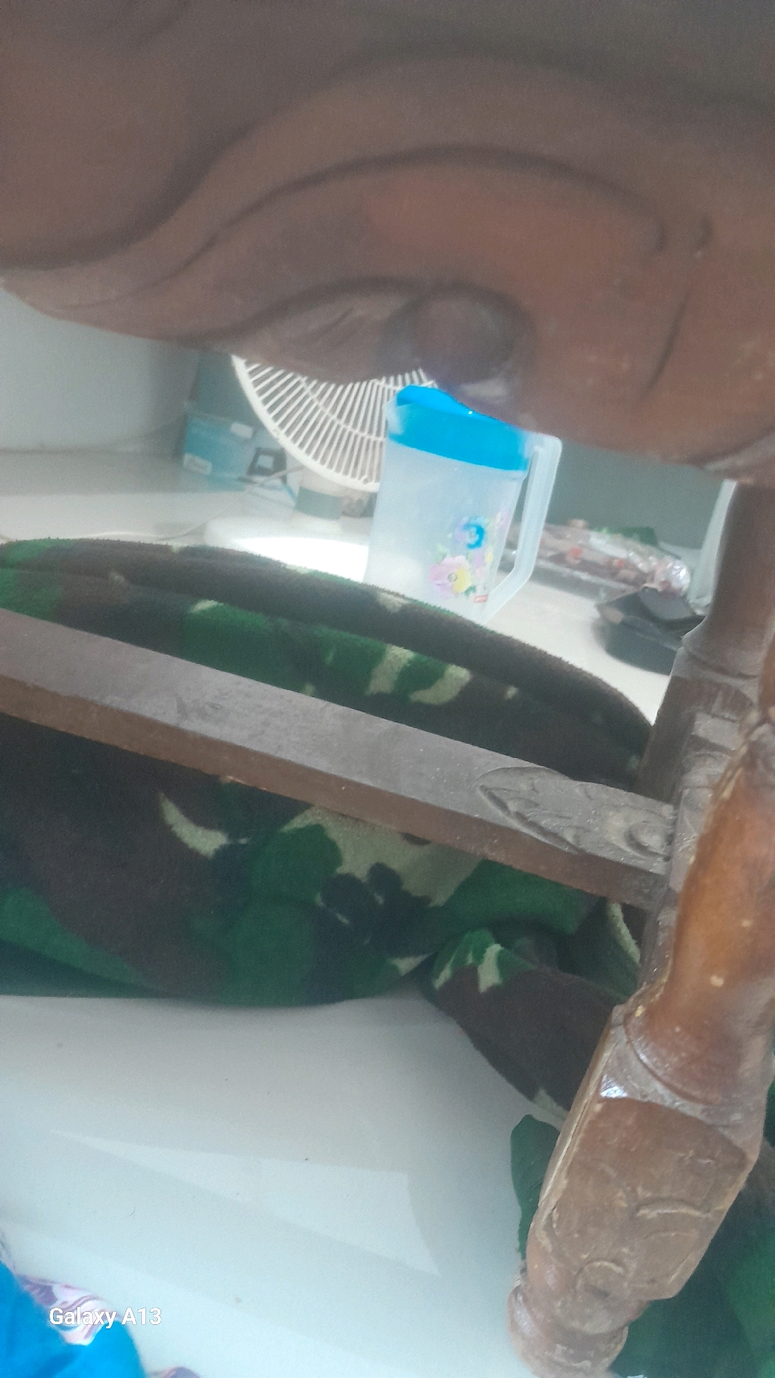 The image appears to be taken from a low angle looking upwards at the underside of a piece of dark wood furniture. A white fan with visible blades is partially obscured by the furniture and is positioned behind a clear plastic cup with a blue lid. The cup has a floral design on it. In the foreground, a blanket with a camouflage pattern of green, brown, and black is draped. A thin, flat piece of wood runs horizontally across the blanket. To the right, a wooden leg with carved detailing is visible. The floor appears to be a light-colored surface, possibly tile or linoleum. The text "Galaxy A13" is visible in the bottom left corner, suggesting the image was captured with a Samsung Galaxy A13 phone. The presence of a fan and the general indoor setting suggest this could be a residential or commercial space. Without further context, it is impossible to determine the specific location within Hadipolo, Indonesia.