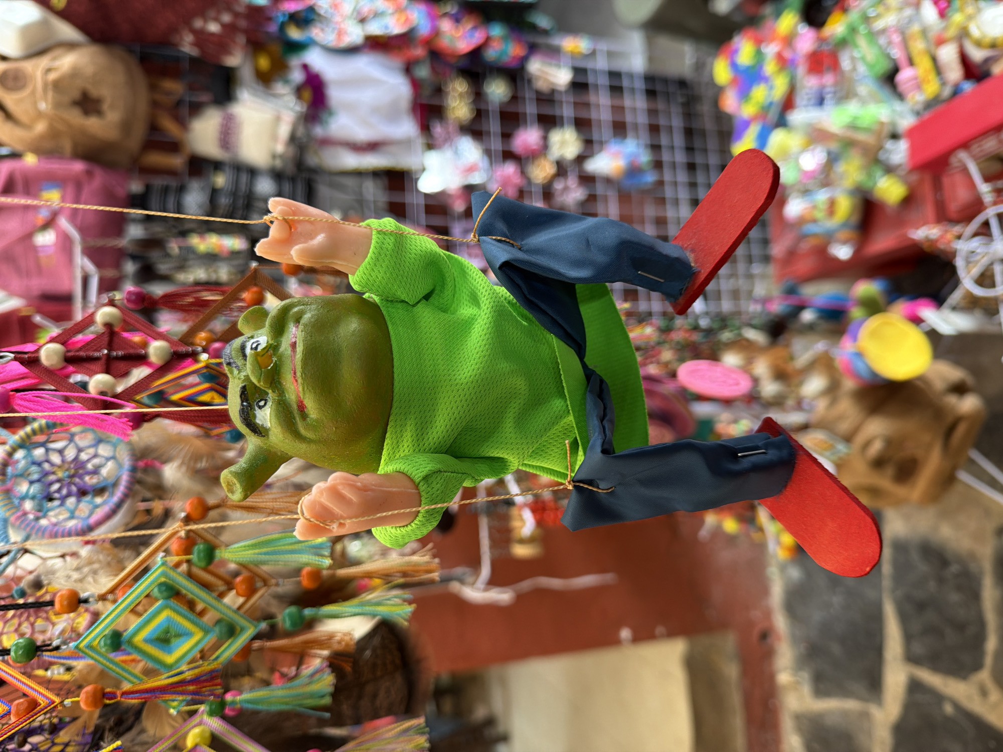 In Cuetzalan, Mexico, a green, frog-like puppet with red shoes hangs from strings amidst an array of colorful, handcrafted items. The puppet, dressed in a green shirt and dark blue pants, is suspended mid-air with its arms and legs outstretched. Surrounding it are vibrant dreamcatchers and other artisanal goods displayed on a wire mesh in a marketplace. The background consists of various items for sale, creating a bustling and visually rich scene. The floor is stone.