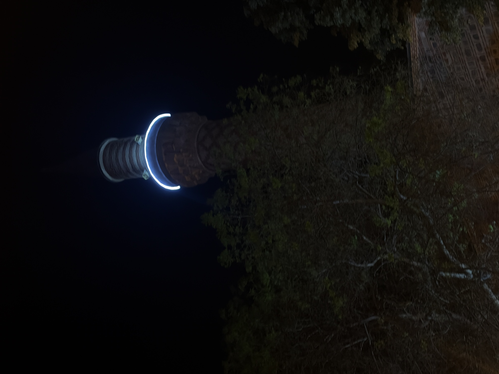 The image shows the Nebet Tepe tower in Plovdiv, Bulgaria, at night. The tower is illuminated by a semi-circular neon light at its top. The structure is partially obscured by trees. The background is dark, indicating a nighttime setting.