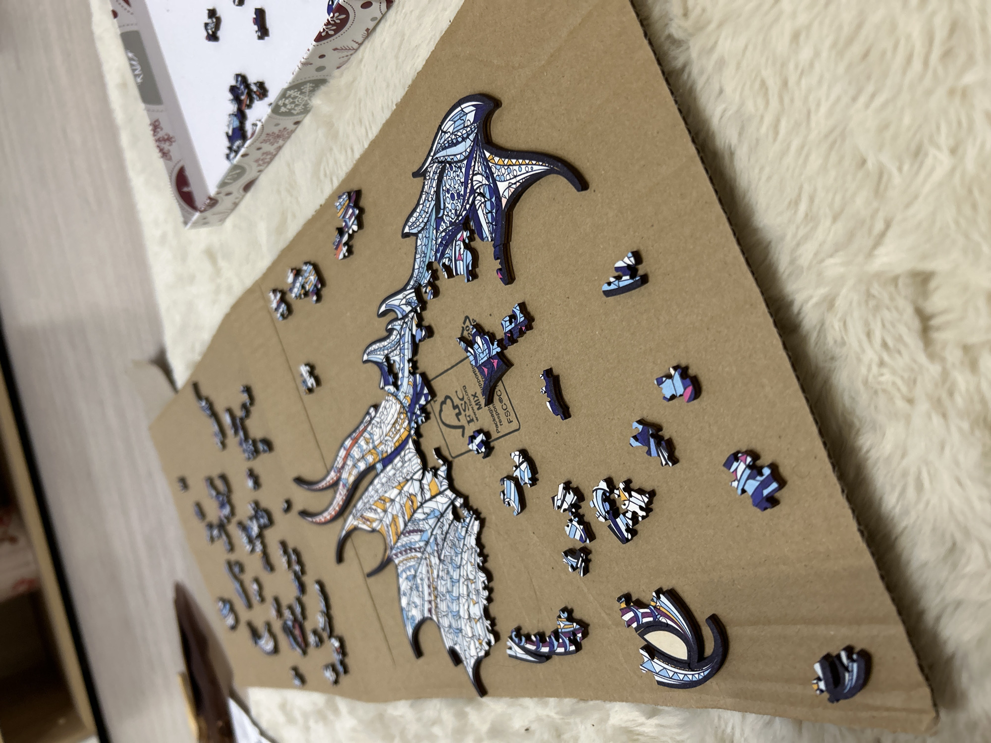 A partially completed wooden jigsaw puzzle lies on a brown cardboard surface against a backdrop of cream-colored, fluffy material, possibly a rug or blanket. The puzzle depicts what appears to be a stylized dragon with intricate patterns and colors, mainly shades of blue, white, and yellow. Several puzzle pieces are still detached and scattered around the partially assembled dragon wings. The puzzle box is visible in the upper left corner, holding more unassembled pieces. The scene is in a location with artificial light. There is no clear evidence that this image was taken in the city of Varshets, Bulgaria.