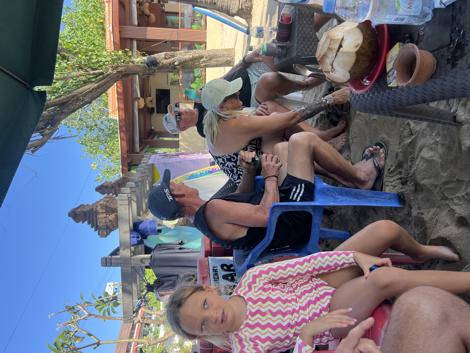 A group of people are gathered around a table on a sandy beach in Kuta, Indonesia. The scene is outdoors under a large umbrella, with a clear blue sky. In the foreground, a young girl with blonde hair, wearing a pink and orange striped long-sleeved rash guard, is seated on a red chair. To her right, a man in a black sleeveless shirt and shorts is sitting on a blue chair. Behind them, a woman in a black and white patterned top and a cap is leaning forward. Another man, wearing a cap and sunglasses, is seated further back.
On the table in front of them, there is a halved coconut, a bottle of Sprite, a bottle of water, a pack of cigarettes, and a small brown pot. Several surfboards are visible leaning against a structure in the background, suggesting a surf-related setting. Clothing items are hanging on a rack behind the people, and a wooden structure with intricate carvings is also present. The overall atmosphere appears relaxed, with the group likely enjoying refreshments on the beach.