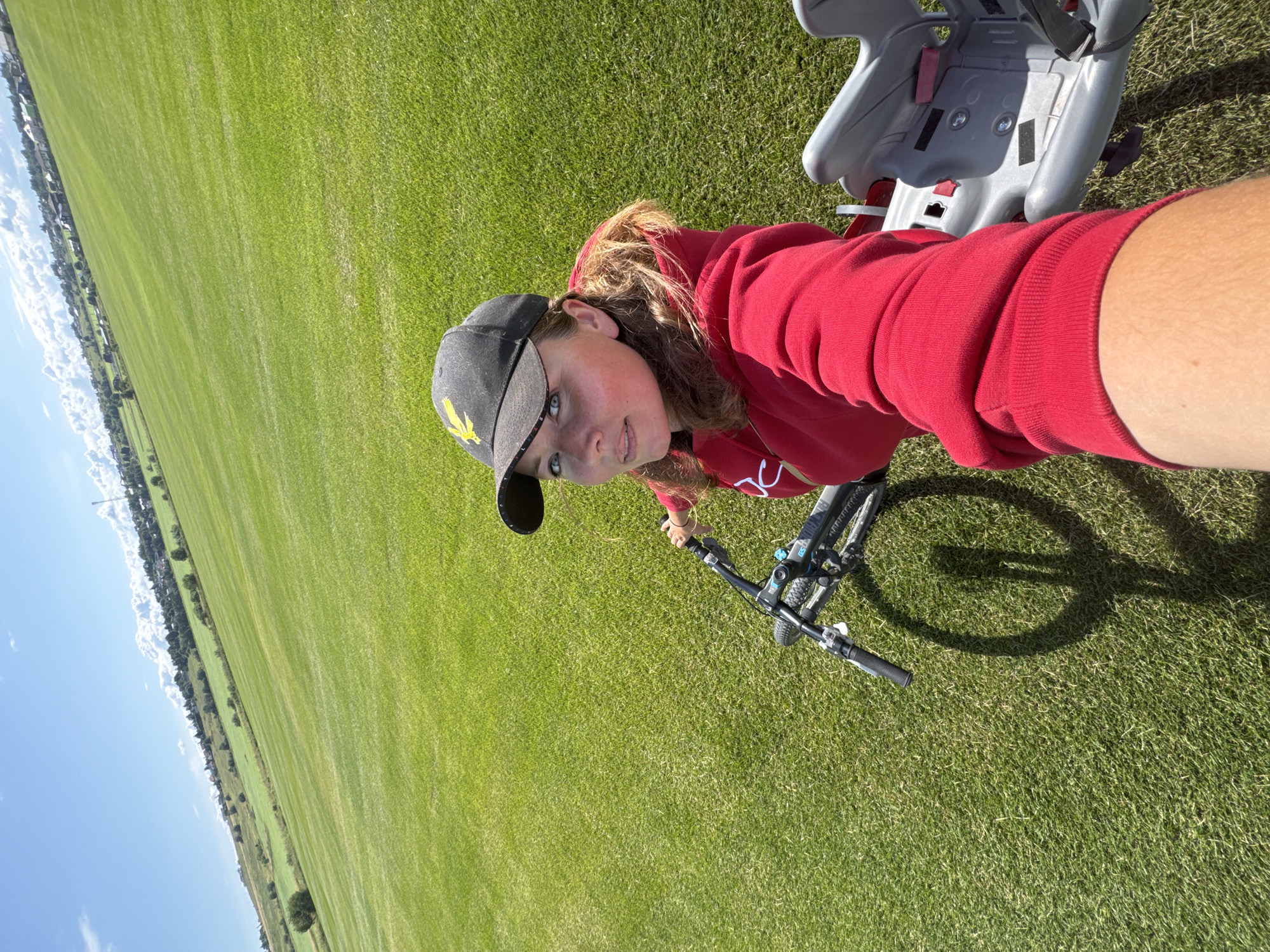 A woman is taking a selfie while sitting on a bicycle with a child seat. She is wearing a red hooded sweatshirt and a black baseball cap with a yellow emblem. The bicycle is on a lush green grassy field. In the background, there is a gentle slope covered in grass, with some trees and buildings on the horizon. The sky is blue with some scattered white clouds. The scene appears to be in an outdoor recreational area.