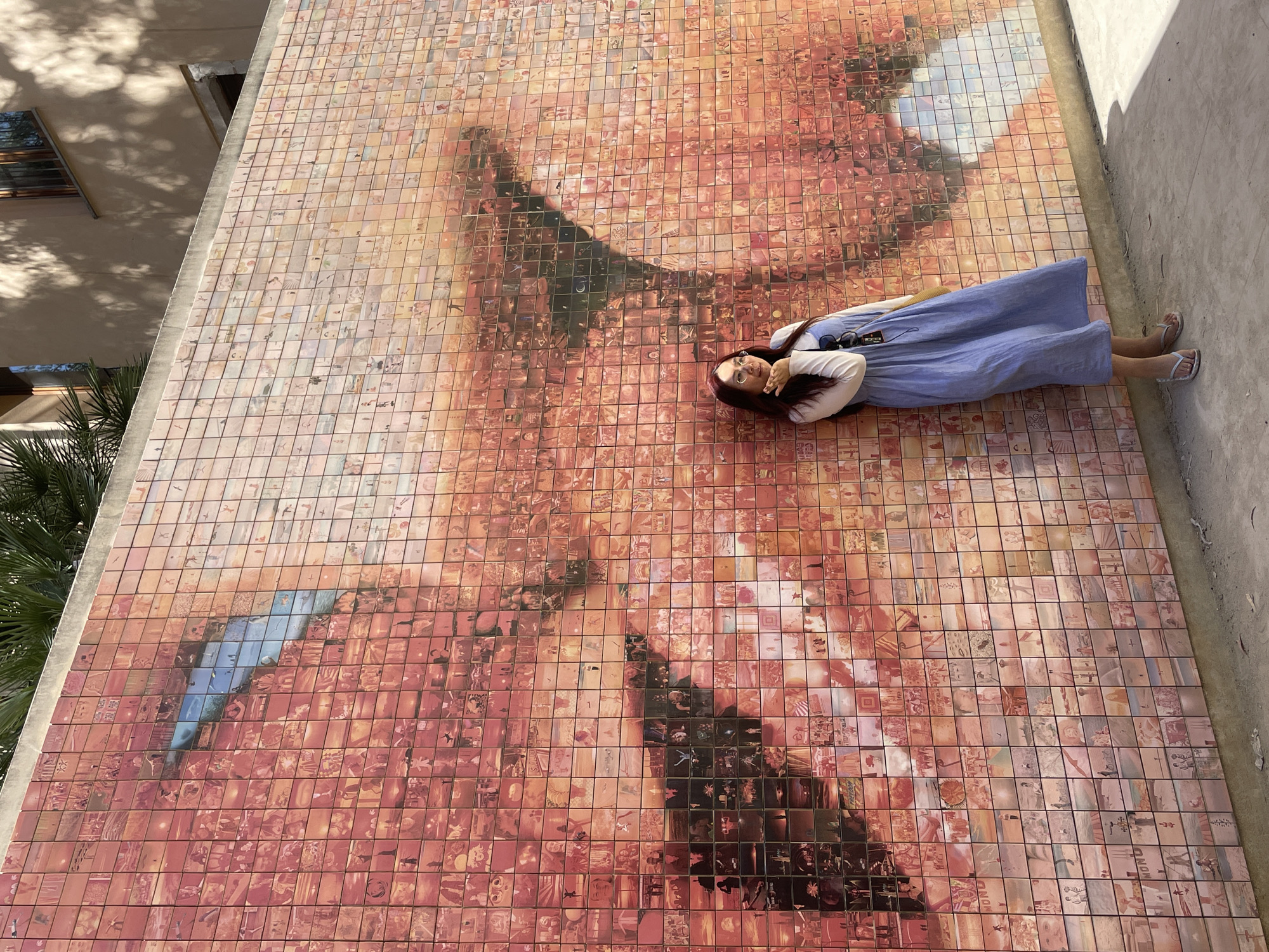 A young woman leans against a massive mosaic mural in Barcelona, Spain. The mural is composed of numerous small tiles, each depicting a scene or image, that together form a larger portrait. The woman, wearing a blue dress and a white cardigan, has dark hair and is looking away from the camera with her hand near her face. The mural covers a large portion of a building's exterior wall, with palm fronds visible at the top. The ground in front of the wall is paved, and shadows from trees are cast across the scene. The overall impression is one of vibrant urban art juxtaposed with everyday life.