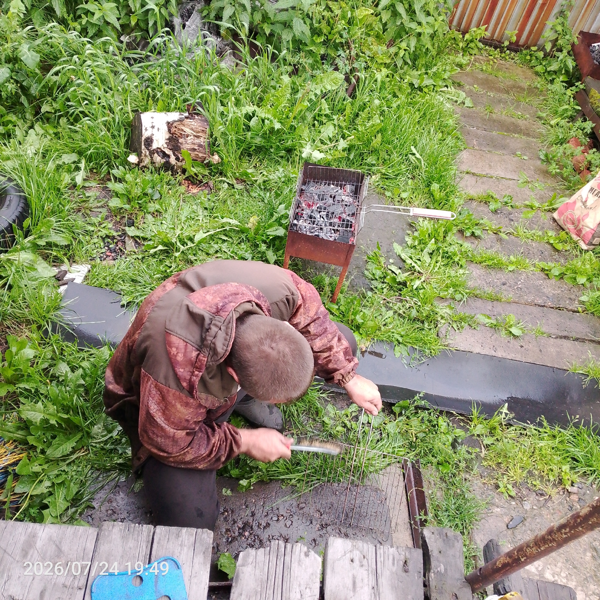 In an outdoor setting with abundant green grass and vegetation, a person wearing a camouflage jacket and dark pants is cleaning a metal grill. The person is crouching and using a brush to scrub the grill rack. Nearby, a portable barbecue grill with hot coals is visible. The ground appears wet and muddy. Wooden planks form a pathway or platform in the foreground, and a portion of a metal fence can be seen in the background. A bag of charcoal briquettes is also present. The scene is captured from a high-angle perspective.