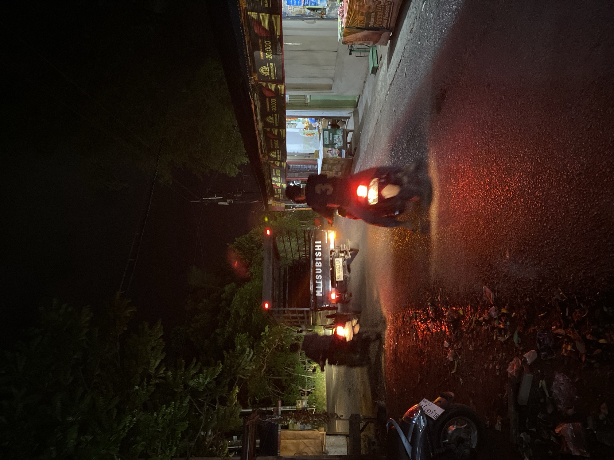 In Aneuk Paya, Indonesia, at night, a street scene unfolds with limited visibility due to darkness. The road surface is wet, reflecting the red and white lights from vehicles and possibly overhead streetlights. To the left, a parked motorcycle is partially visible. Further down the road, a Mitsubishi truck with the brand name prominently displayed travels away from the viewer. The truck has a flatbed and appears to be carrying cargo. In front of the truck, a motorcyclist is riding away from the viewer, their silhouette visible against the vehicle's taillights. The motorcyclist is wearing a dark shirt with a white number "3" on the back. Along the right side of the road, a row of shops or stalls with awnings are present, with some lights illuminating their interiors. Banners with text, some indicating prices, are hung above the stalls. The environment is lined with trees, their foliage dark against the night sky. Trash is scattered on the wet ground near the parked motorcycle on the left.