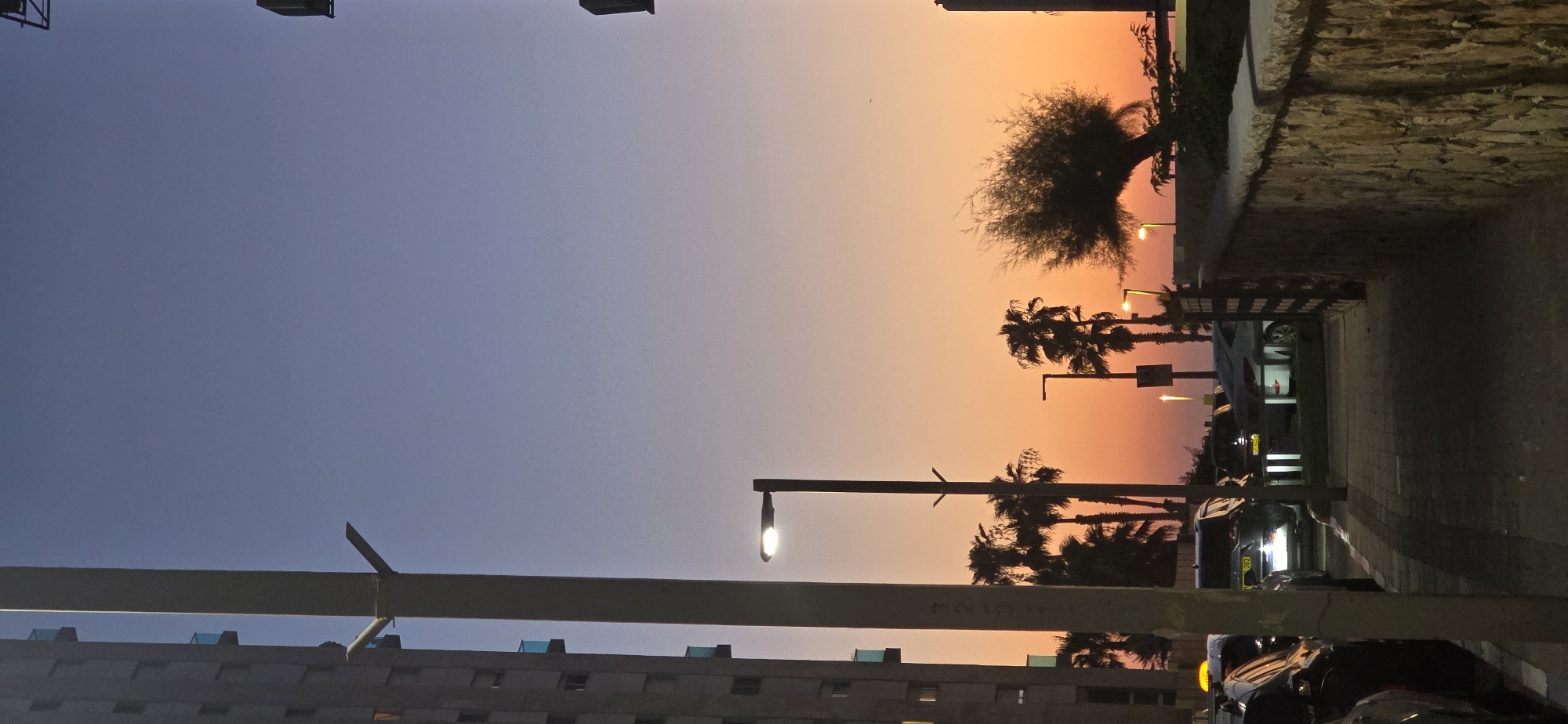The image depicts a street scene in Bat Yam, Israel, at dusk. A tall, modern building is visible on the left side of the frame. A lamppost, illuminated, stands in the foreground. The sky exhibits a gradient of colors, transitioning from a pale blue at the top to warm orange and yellow hues near the horizon. Several palm trees are scattered throughout the scene, silhouetted against the colorful sky. The street appears to be lined with parked cars. A stone wall runs along the right side of the image, bordering a pathway. The overall impression is one of a coastal urban environment as day transitions into night.