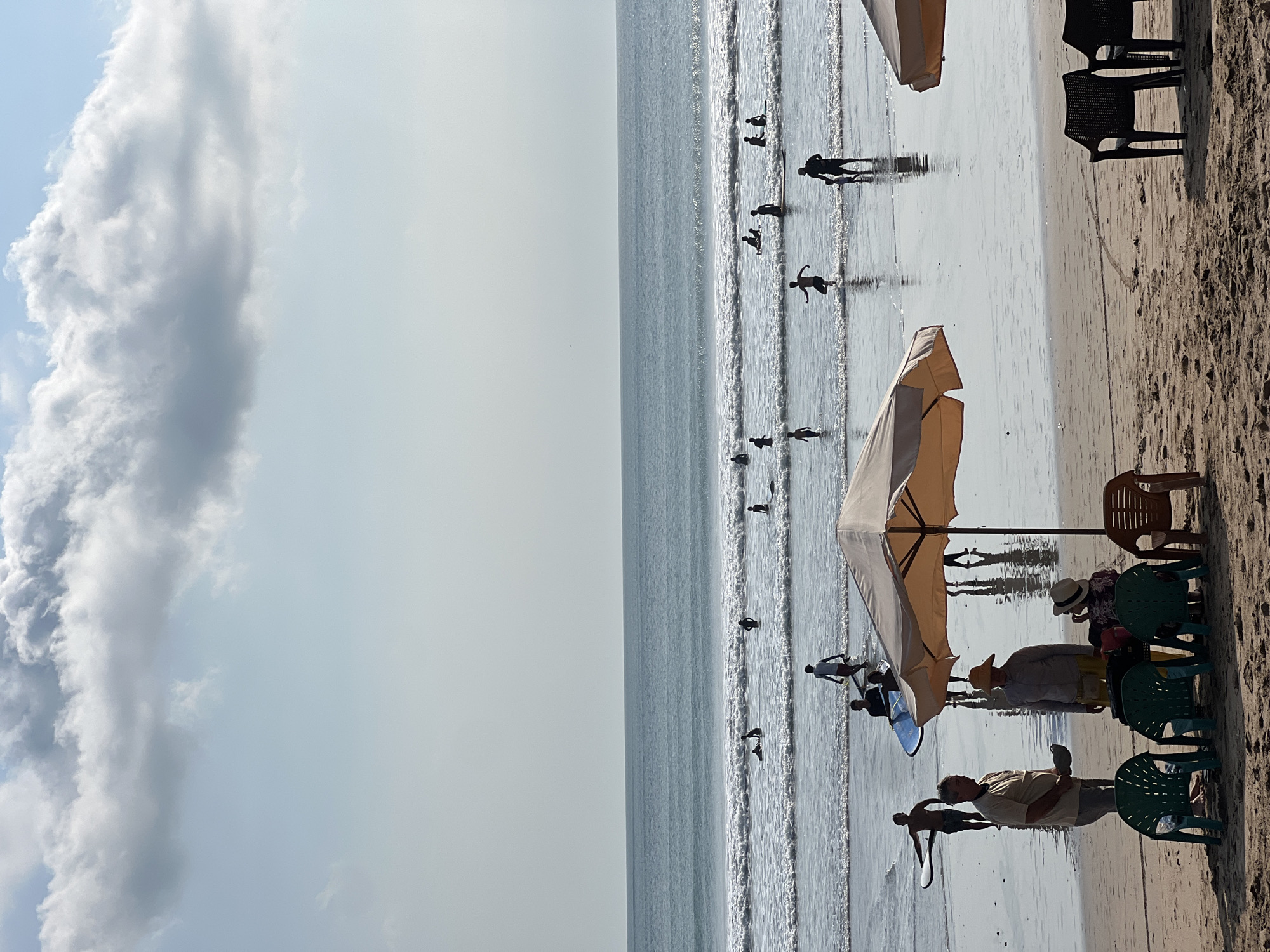 The image depicts a lively beach scene in Kuta, Indonesia, likely during the daytime given the bright sky and sunlight. In the foreground, two large beach umbrellas offer shade over empty chairs on the sandy shore. Closer to the water, a group of people are visible. Two men stand talking, one holding a hat. Another person wears a wide-brimmed hat and sits facing away from the camera. Further back, more people are engaged in beach activities. Several individuals, some carrying surfboards, are in the shallow water, either wading or preparing to surf. Others are further out in the ocean, with some appearing to be riding waves or waiting for them. The ocean itself is a calm blue, with gentle waves breaking on the shore. The sky above is a pale blue with scattered, fluffy white clouds. The overall atmosphere is relaxed and recreational, indicative of a popular tourist destination.