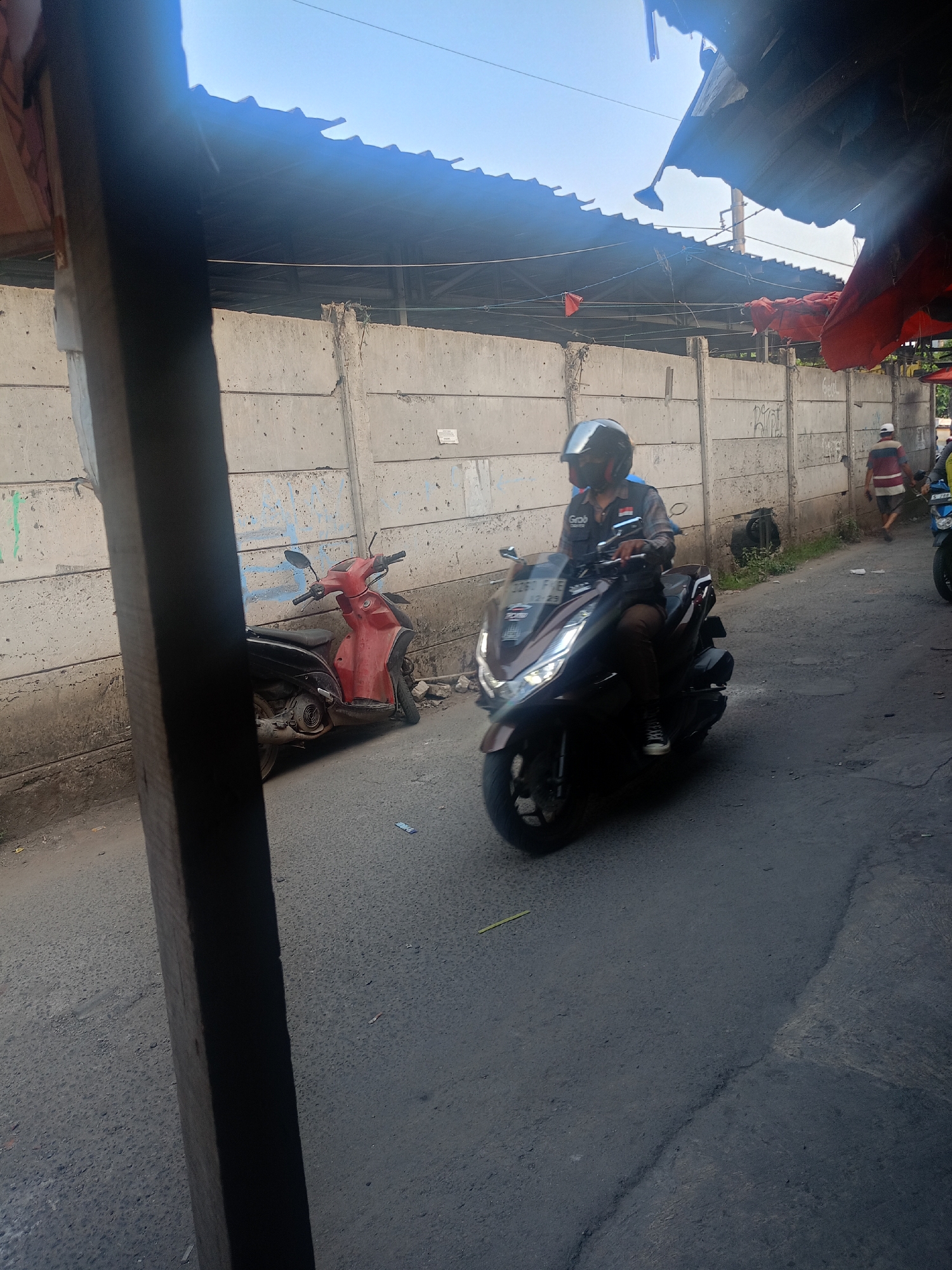 In Bojong Gede, Indonesia, a scene unfolds along a dusty, paved road adjacent to a concrete barrier wall. The wall, marked with graffiti, appears to be part of a market or industrial area, with a corrugated metal roof structure partially obscuring the sky overhead. On the road, a man wearing a helmet rides a dark brown scooter, identified by a "Grab" logo on his jacket. He is positioned in the center of the frame, moving away from the camera. To the left, parked on the side of the road, is a red and black scooter. Further down the road, a man in a striped shirt walks away from the camera, and another scooter is partially visible. The lighting suggests it is daytime, with clear skies.