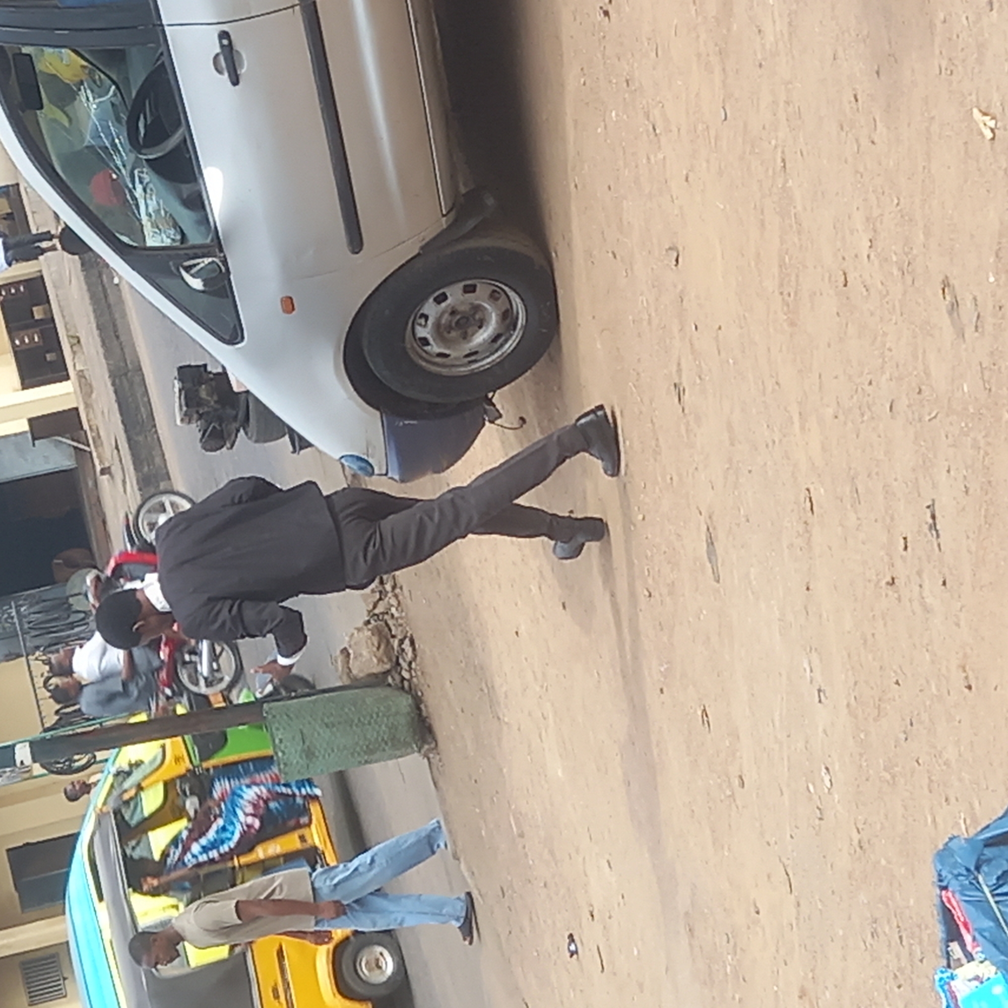This image captures a vibrant street scene in Sardauna, Nigeria, during bright daylight. The ground appears dusty and unpaved in places, typical of local roads.
In the foreground, a white compact car, possibly a hatchback, is prominently parked on the left. Its windshield shows significant damage, appearing shattered or extensively cracked. Adjacent to the car, a man dressed in a black suit with black trousers and black dress shoes is visible. He is positioned as if walking or leaning, facing away from the camera and slightly to the left, towards the car or something on the ground near it.
Further down the street, behind the suited man, a red and black motorcycle is parked. Several other individuals are scattered throughout the scene. To the right, a distinctive yellow and blue auto-rickshaw, commonly known as a "keke Napep" in Nigeria, is present. People are visible inside and around the keke Napep, including one person covered in a brightly patterned, blue and white cloth, and another man in a light t-shirt and blue jeans walking alongside it.
The background features various buildings lining the street, suggesting a commercial or residential area. The overall impression is that of a bustling local thoroughfare on a clear, sunny day. No discernible text is visible on any objects or signs within the frame.