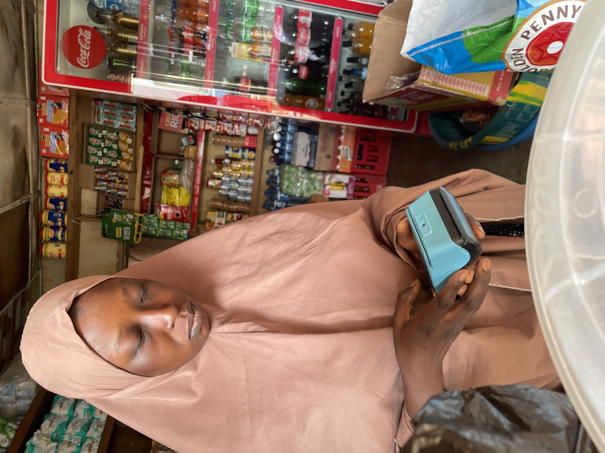 The photo shows a woman wearing a peach-colored hijab standing in what appears to be a small store or shop. She is holding a light blue point-of-sale (POS) terminal in her hands. The background of the image shows a variety of items commonly found in a shop, including a refrigerator stocked with beverages like Coca-Cola, shelves with packaged foods and other consumer goods. A "PENNY" branded bag is visible on top of the cooler. The location of the shop is speculated to be in Jos, Nigeria.