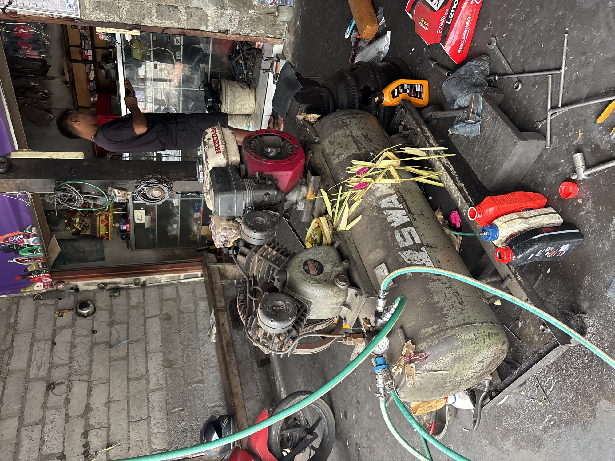 The image depicts an outdoor workshop or garage in Kuta Selatan, Indonesia, likely a motorcycle repair shop. A large, old, green air compressor with a Honda engine is the central focus. It has a visible gauge and is connected to green hoses. To the left, the front wheel and part of a red motorcycle are visible. A black motorcycle helmet rests on a wooden bench behind the motorcycle.
In the background, a man in a black t-shirt and dark pants stands near shelves filled with various items, possibly motorcycle parts or supplies. The workshop walls are constructed from grey cinder blocks. The ground is a dark, possibly concrete, surface, with visible oil or water stains and scattered tools. Various bottles of liquids, including engine oil, are placed around the air compressor. Other items such as tires, a hammer, and wrenches are also present in the scene. The overall environment appears utilitarian and well-used, consistent with an active repair shop.