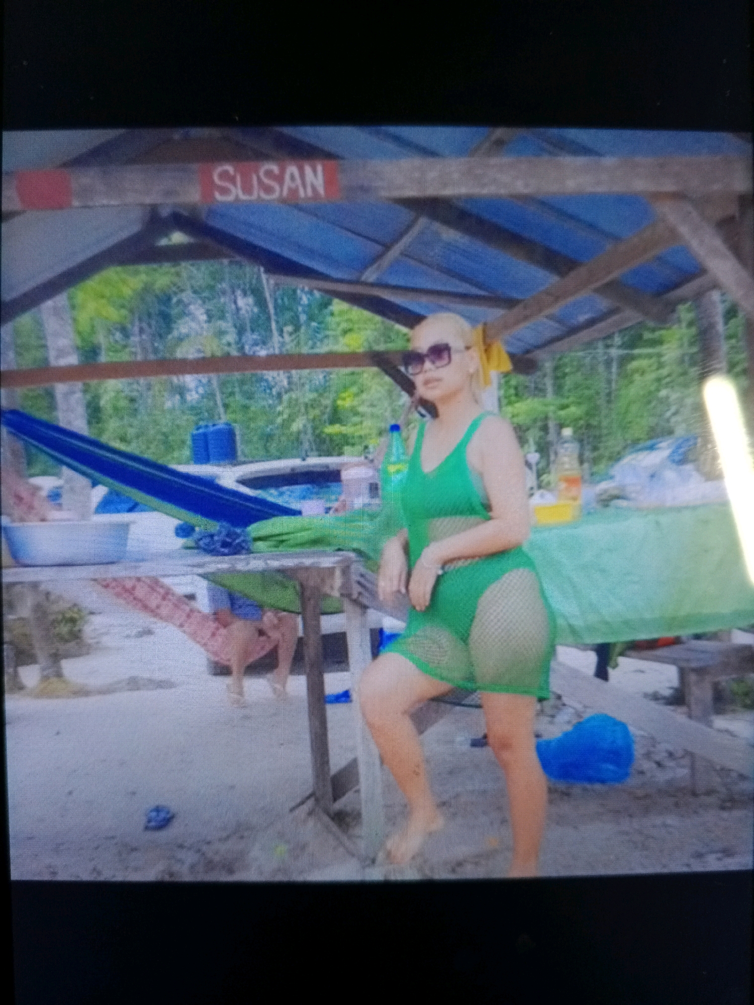 A woman wearing a green mesh bikini and sunglasses stands in a sandy outdoor area, possibly a beach or a tropical setting. She has blonde hair and is posing with one hand on her hip. In the background, there is a blue hammock strung between two poles, a table covered with a green cloth, and a structure with a blue tarp roof. A sign with the word "SUSAN" is visible above the structure. The environment appears to be in Noord, Suriname.