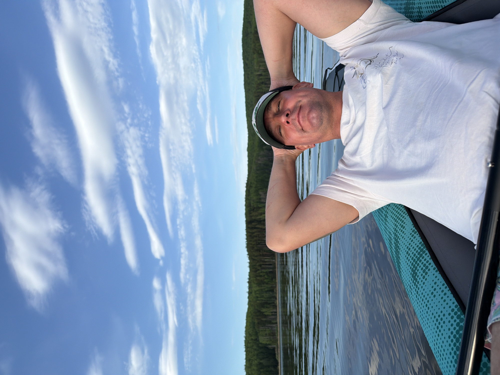 A medium shot depicts a man lying on his back on what appears to be a paddleboard in the city of Chaykovsky, Russia. The man is fair-skinned, wearing a white t-shirt and a cap. His arms are raised, with his hands behind his head. The scene shows water reflecting the sky, with a line of trees visible on the horizon. The sky is blue with scattered white clouds. The paddleboard is turquoise with a black trim. A paddle is also visible, partly out of frame.