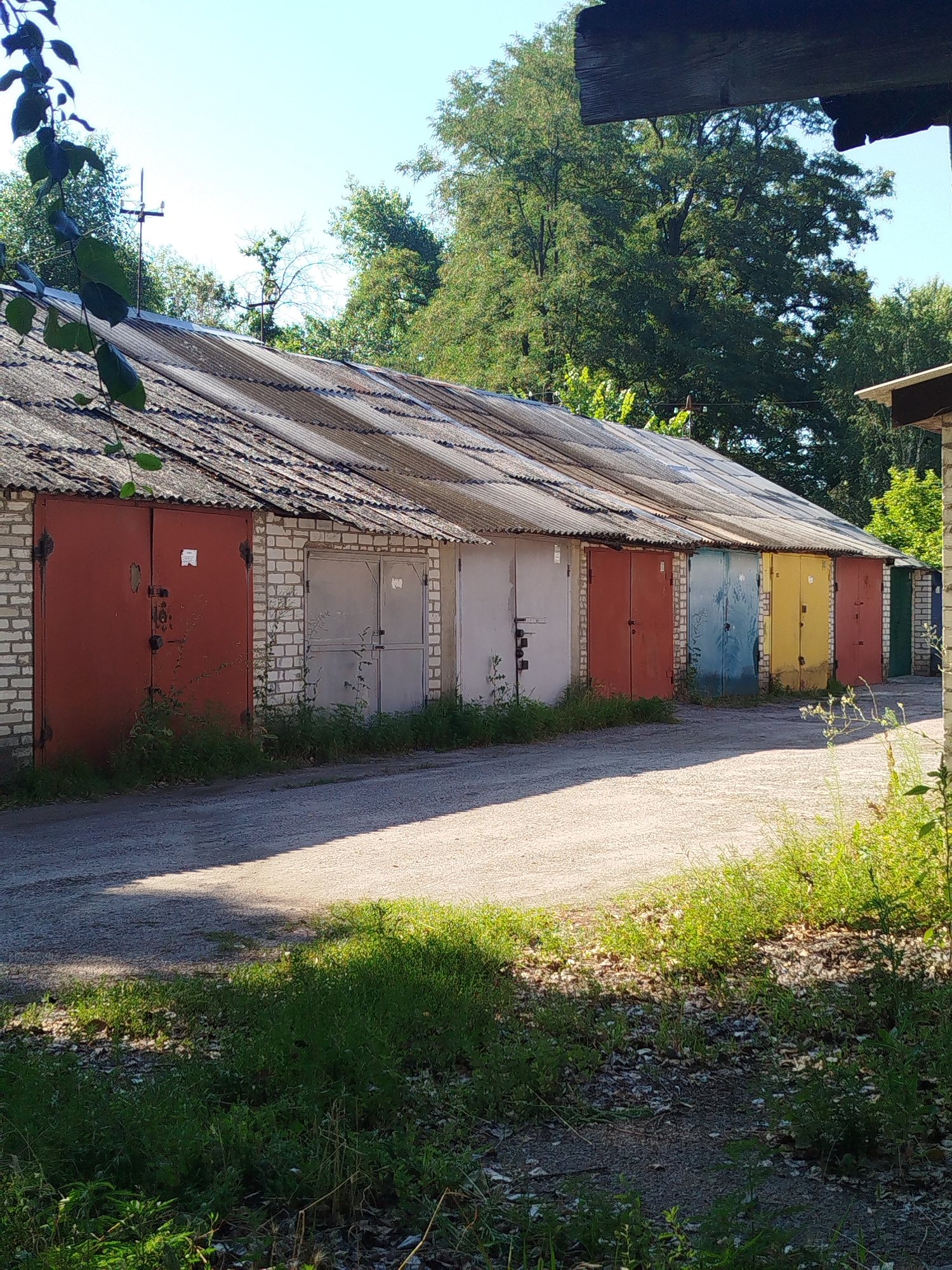 A row of garage doors, painted in various colors including red, gray, blue, yellow, and green, are visible in this outdoor scene. The garages are constructed of brick and have corrugated metal roofs. The ground in front of the garages is a gravel road, and there is overgrown grass and foliage in the foreground and along the sides of the garages. Trees and foliage are visible behind the garages, and the sky is clear blue. The image appears to have been taken on a sunny day. The scene depicts a typical urban or suburban environment, possibly a garage complex. No people or animals are visible in the image.