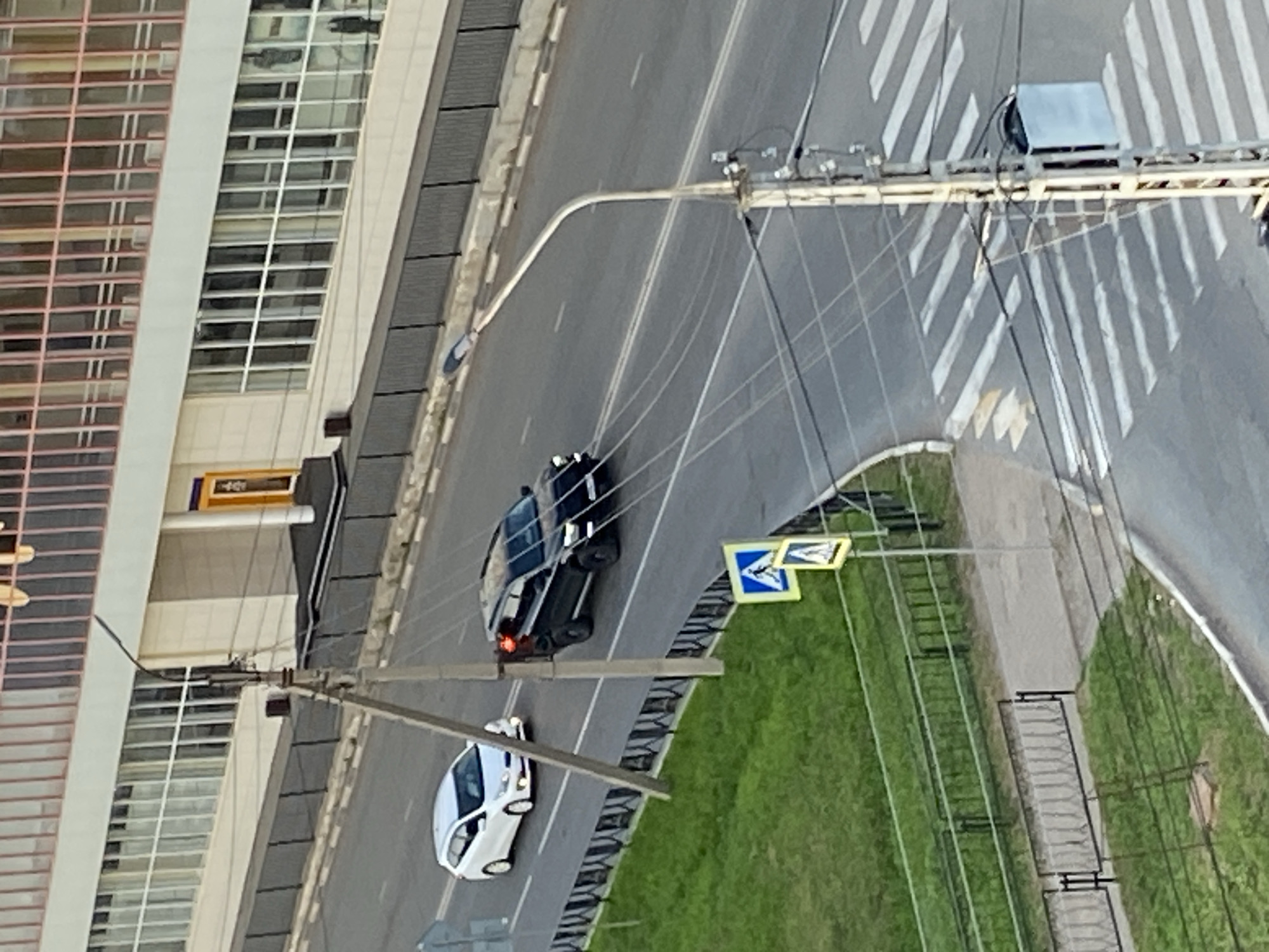 The image depicts a street scene in Neftekamsk, Russia. A black SUV is driving on the road, with its brake lights illuminated. A white hatchback is also visible on the road, ahead of the SUV. A pedestrian crossing is marked on the asphalt, with white stripes indicating the crosswalk. Traffic signs, including a pedestrian crossing sign, are present on the side of the road. A utility pole with electrical wires runs along the street. In the background, a multi-story building with a facade of windows and vertical pink accents is visible. The area includes a grassy verge and a metal fence. The scene appears to be during daylight hours.