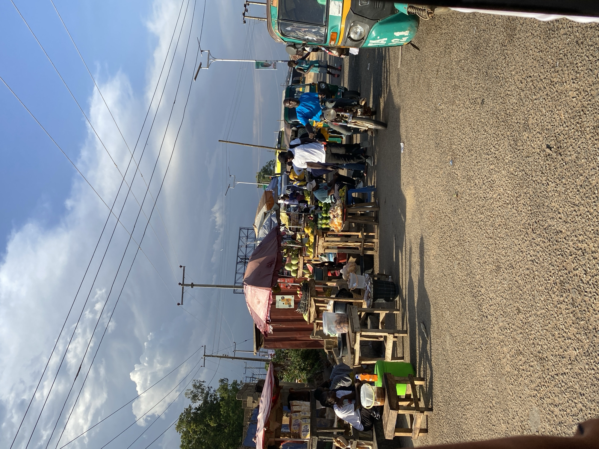 The image captures a street scene in Jos, Nigeria. Along the edge of a paved road, a row of vendors has set up makeshift stalls constructed from wooden pallets and simple tables. These stalls display a variety of goods, including fresh fruits like mangoes and coconuts, along with packaged foods and other merchandise. The stalls are shaded by umbrellas and makeshift canopies. Customers are gathered around the stalls, interacting with the vendors and examining the items for sale. A man in a blue shirt rides a motorcycle on the road, while a green auto-rickshaw is also present. Above the scene, power lines stretch across a sky filled with dramatic clouds. The perspective suggests the photo was taken from an elevated position.