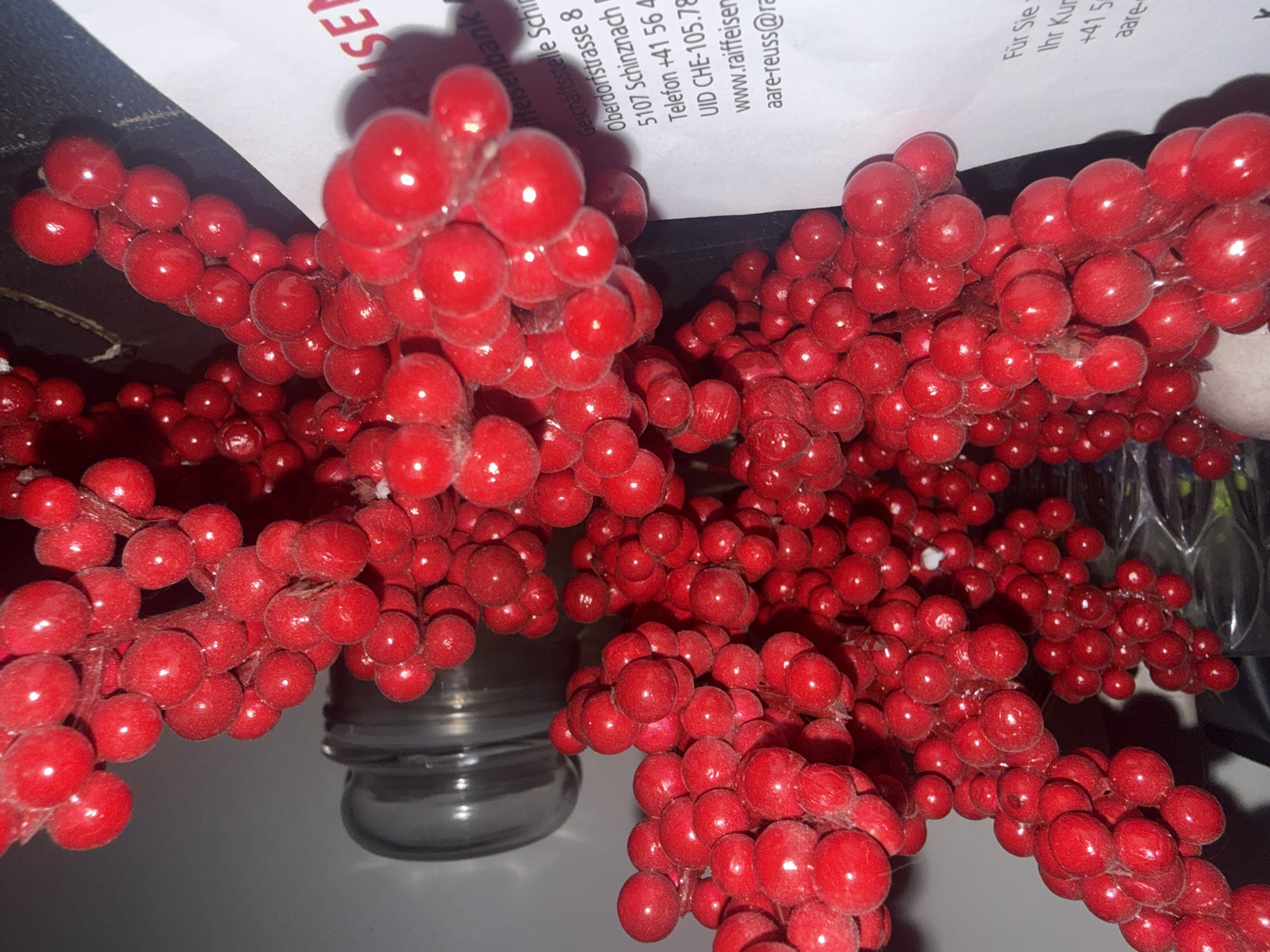 This close-up image captures a vibrant display of red artificial berries, likely part of a decorative arrangement, in Oberflachs, Switzerland. The berries are bright red, round, and appear to have a slightly glossy, possibly flocked or textured surface, giving them a festive or wintery aesthetic. They are densely clustered on thin, wire-like stems, which are mostly obscured by the berries themselves.
In the background and partially beneath the berries, a white document with black text is visible. The text identifies an institution as "iffeisenbank" (Raiffeisenbank), with a "Geschäftsstelle Schi" (branch office) at "Oberdorfstrasse 8" in "5107 Schinznach". Contact information includes "Telefon +41 56 4" and an email address "aare-reuss@ra", along with "UID CHE-105.78" and "www.raiffeisen". Other fragmented text includes "Für Sie" and "Ihr Kur".
A dark background is visible in the upper left, while a glass object, possibly a jar or vase, is partially seen towards the bottom center and right, reflecting some light. No specific activity is depicted; the image is a static shot focusing on the decorative berries and the incidental document. The lighting appears to be artificial indoor light, highlighting the berries' texture and color.